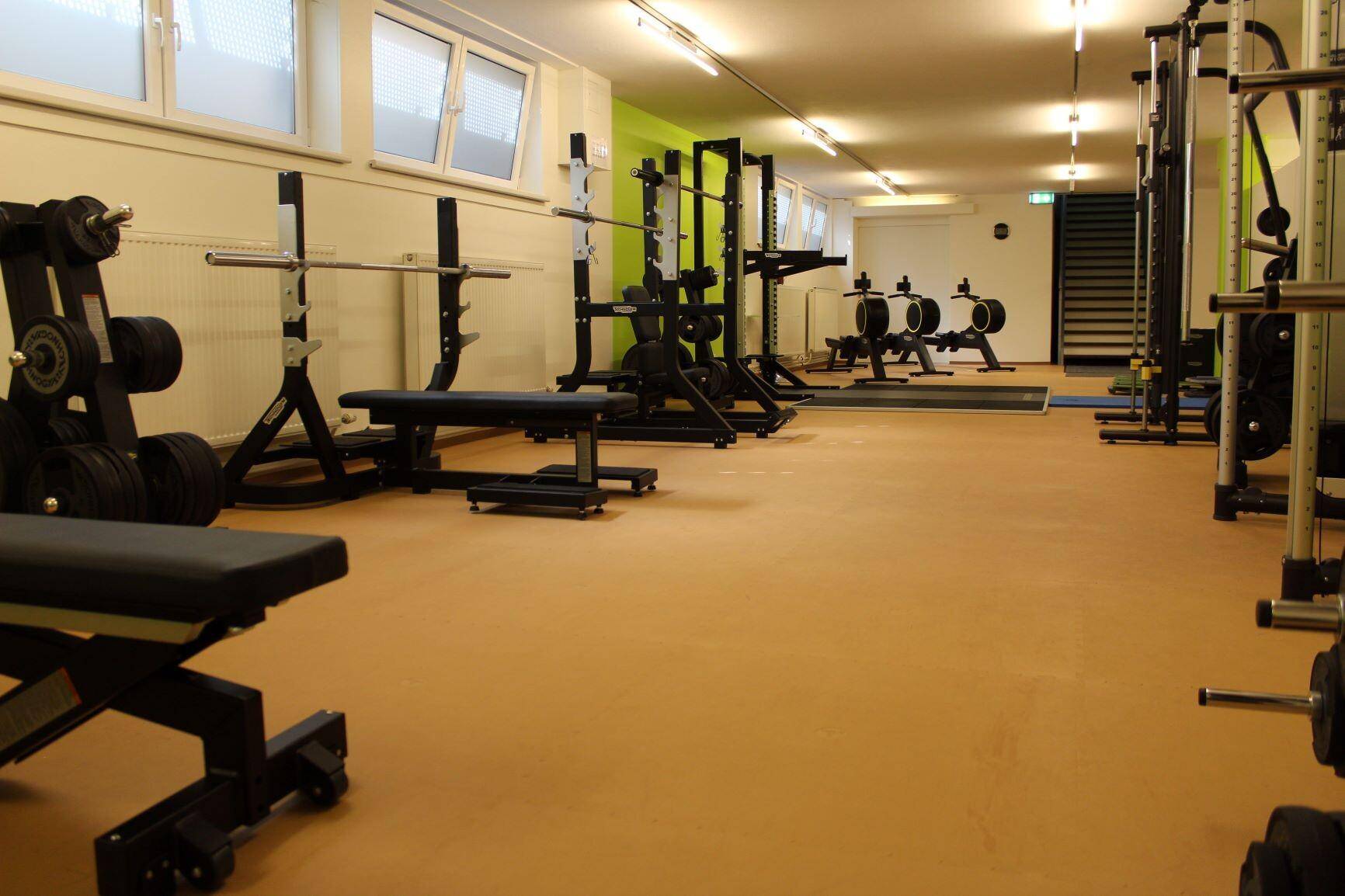 Trainingsbereich mit Hanteln, Bänken und Kraftgeräten unter hellen Fenstern für gute Belüftung.