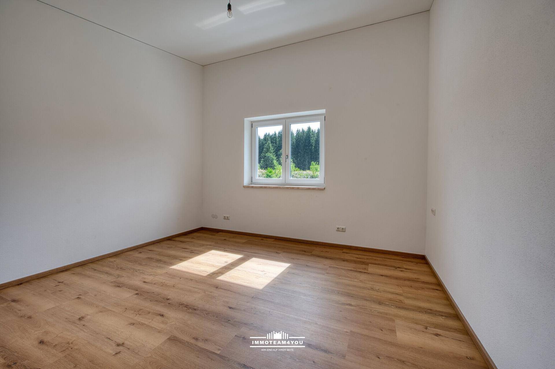 Leeres Schlafzimmer mit Holzboden, weißen Wänden und Fenster mit Blick ins Grüne.