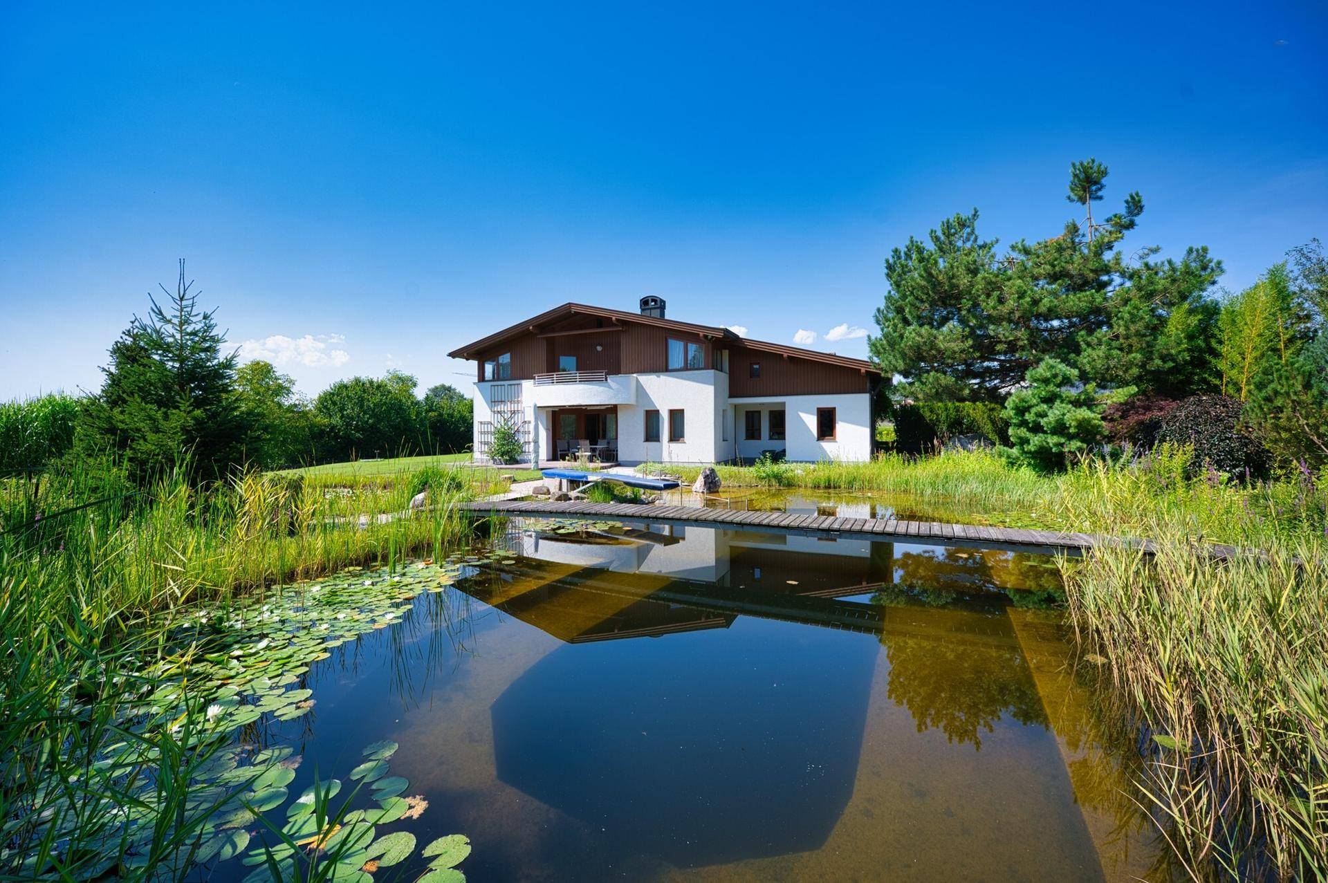 Großzügiges Einfamilienhaus mit Naturteich und weitläufigem Garten unter blauem Himmel.