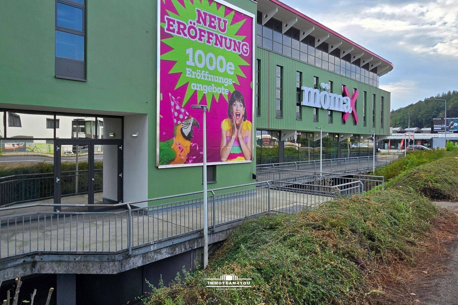 Grünes Geschäftsgebäude mit großer Werbetafel für eine Neueröffnung und einer barrierefreien Rampe.