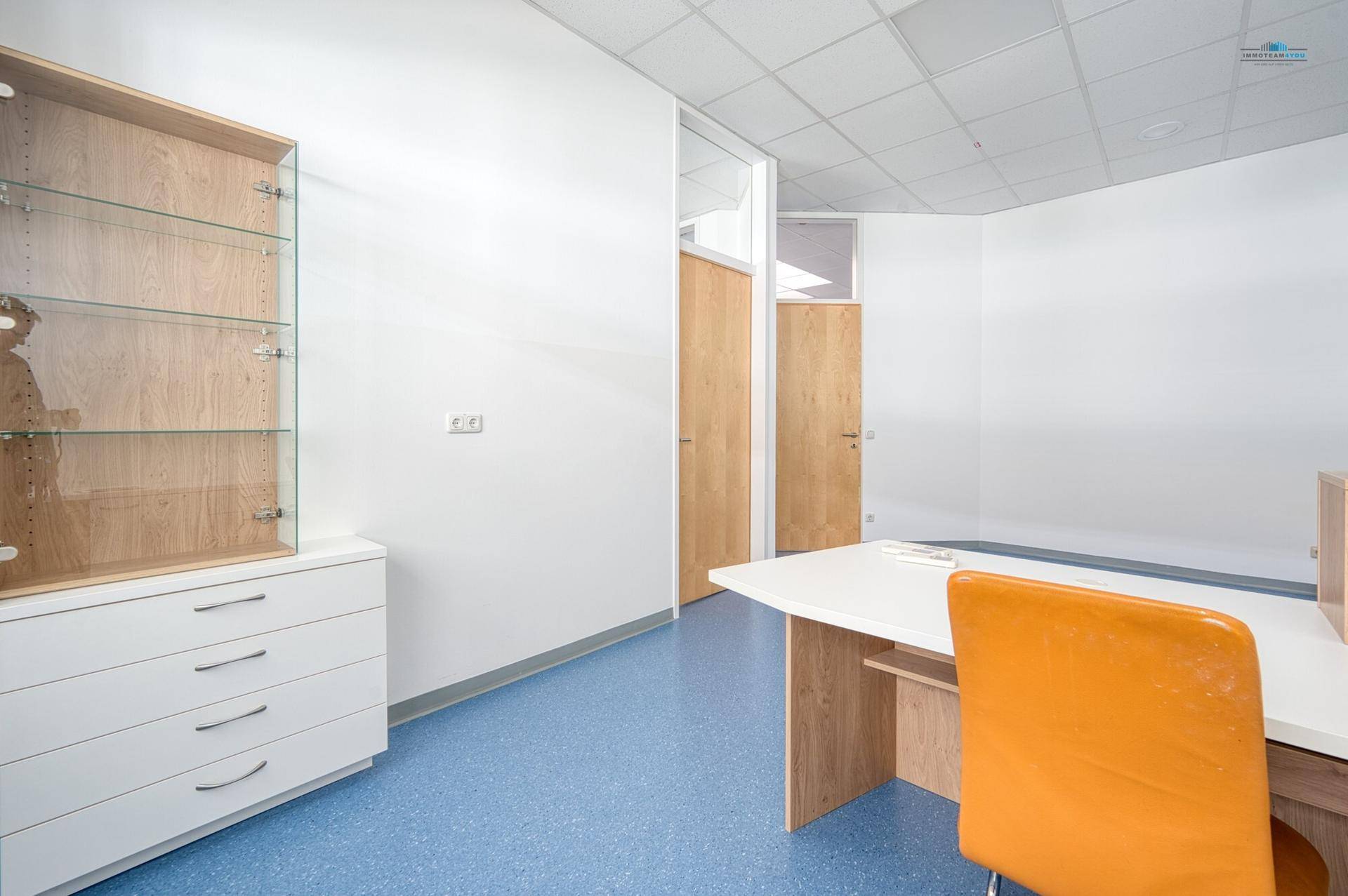 Heller Büroraum mit blauer Bodenfläche, weißen Wänden und funktionalen Möbeln, inklusive Schreibtisch und Schrank.