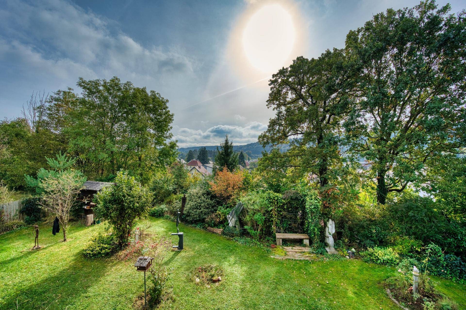 Weitläufiger Gartenbereich mit altem Baumbestand und Sitzgelegenheiten unter strahlendem Himmel.
