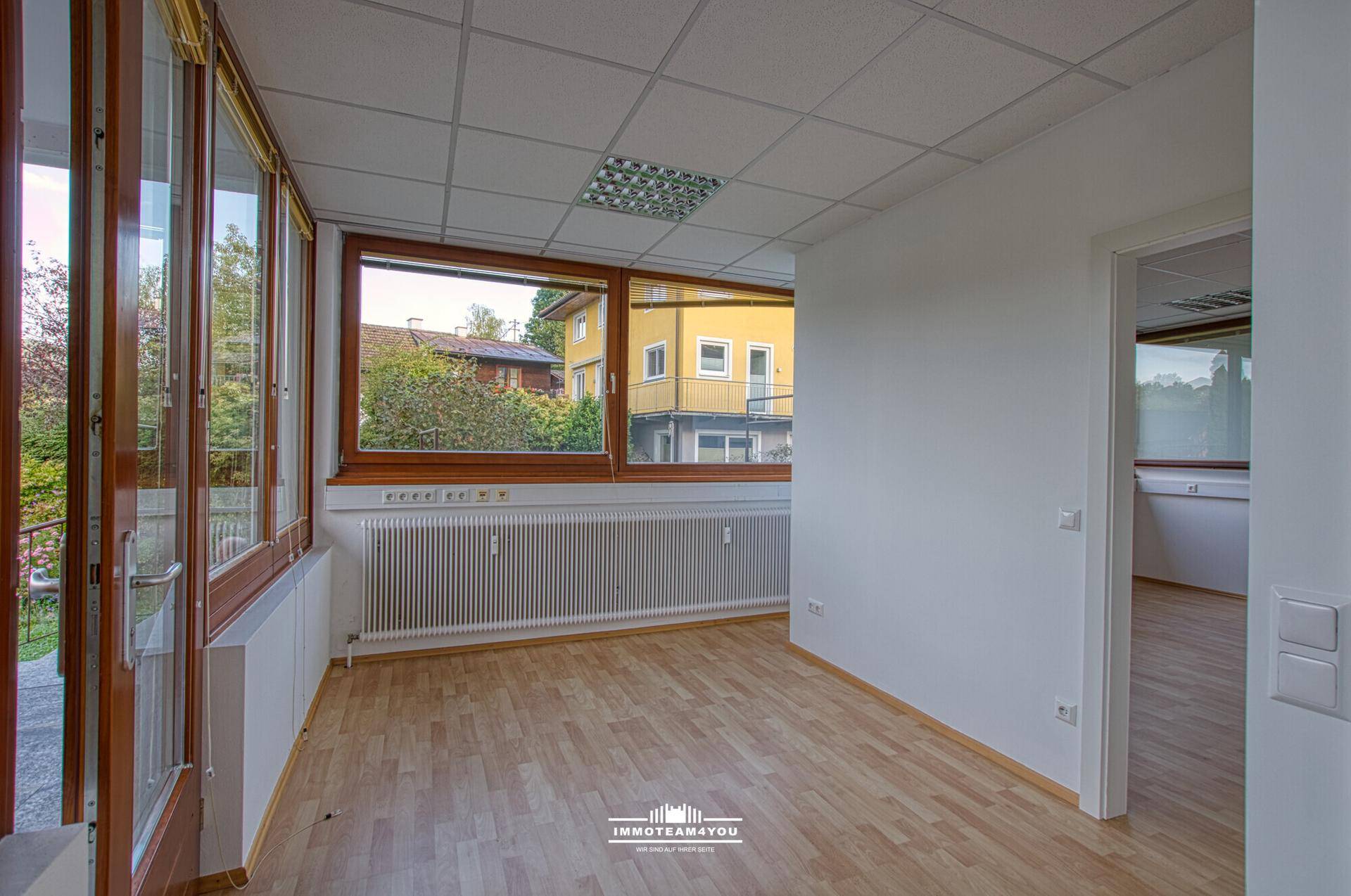 Büroraum mit großen Holzfenstern, Laminatboden und Blick auf die umliegenden Gebäude und Natur.