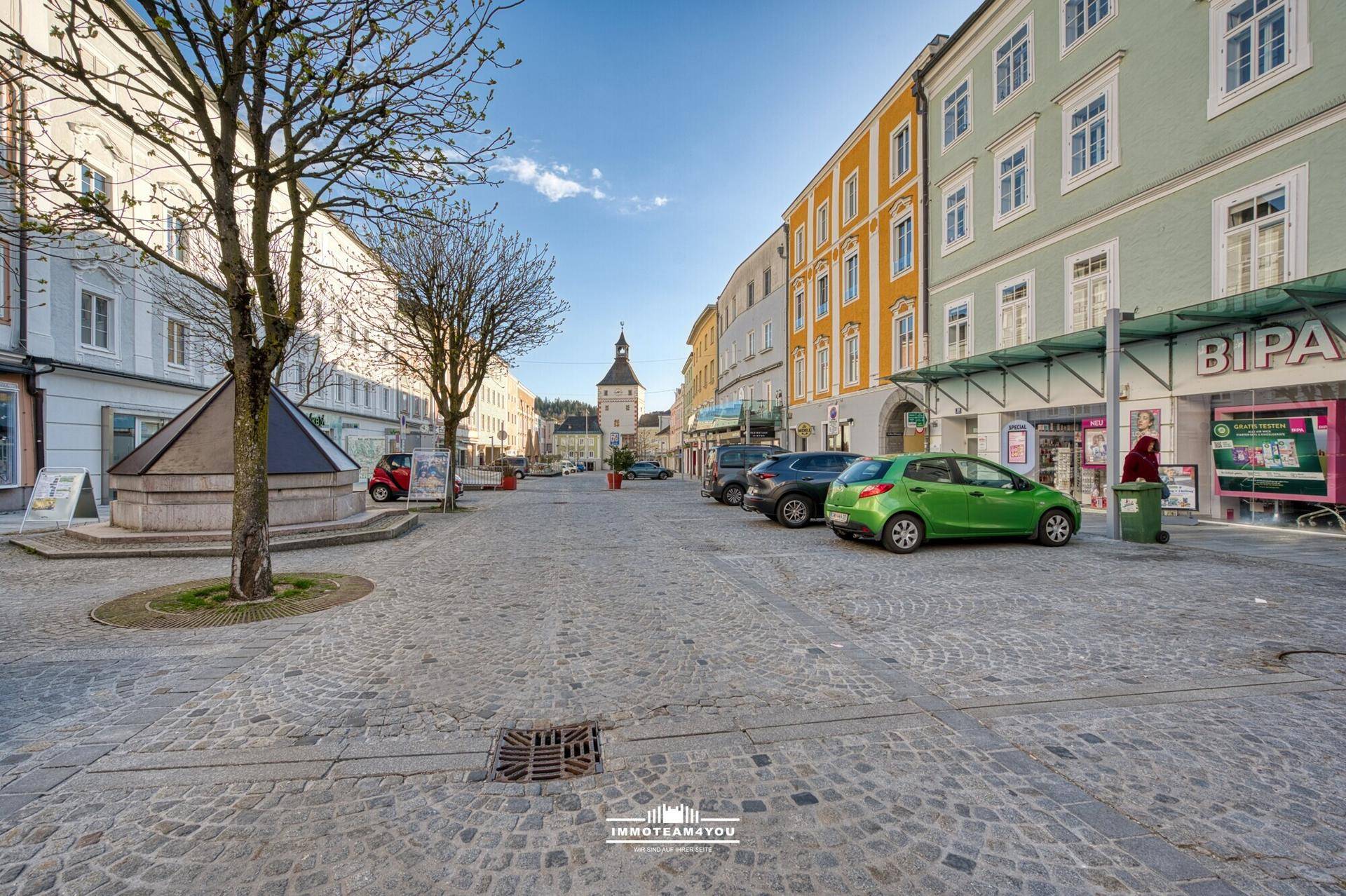 Belebte Stadtstraße mit Kopfsteinpflaster, Geschäften und parkenden Autos unter blauem Himmel.