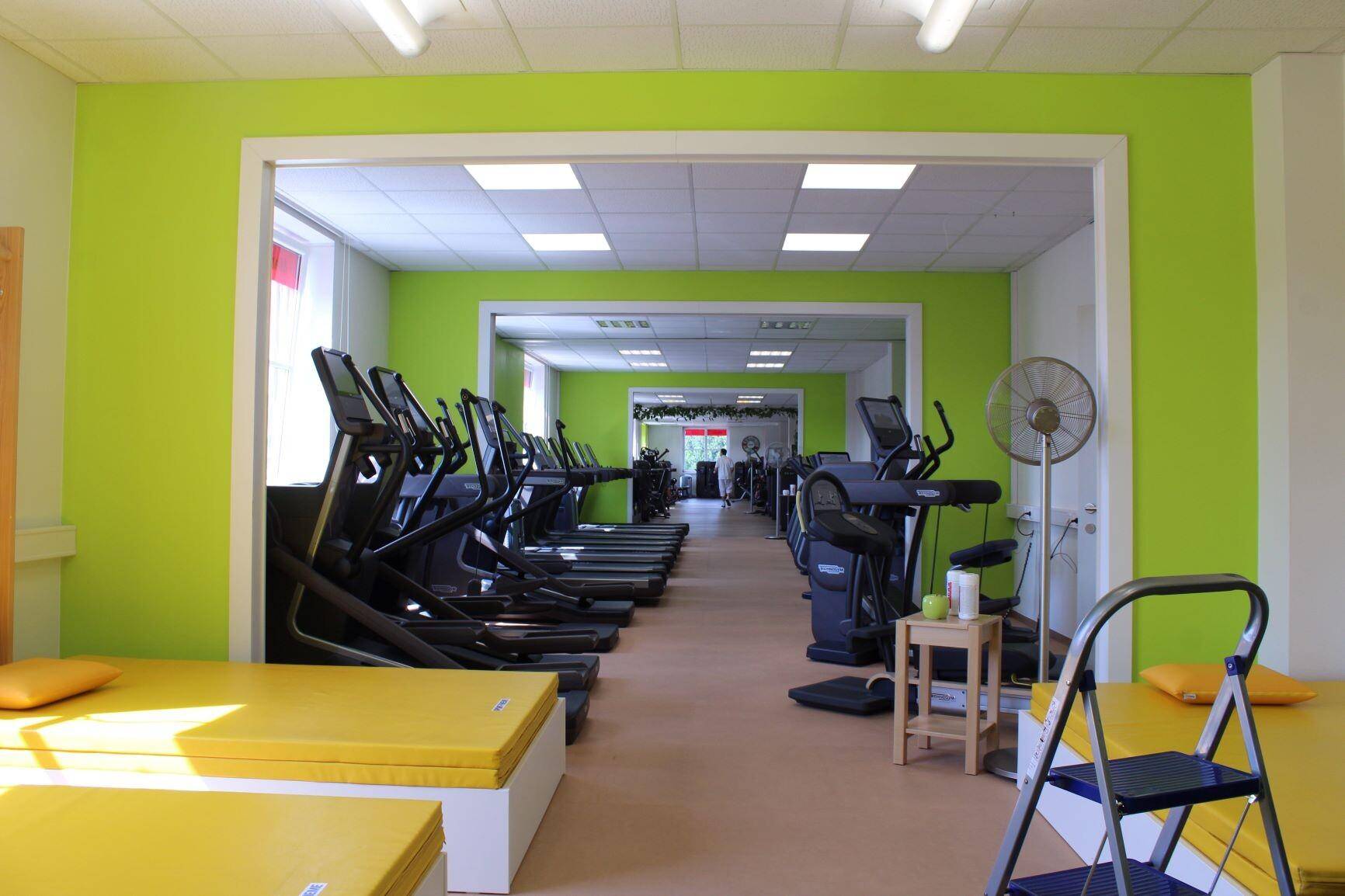Fitnessraum mit zahlreichen Laufbändern und Crosstrainern, Wände in hellem Grün und braunem Boden.