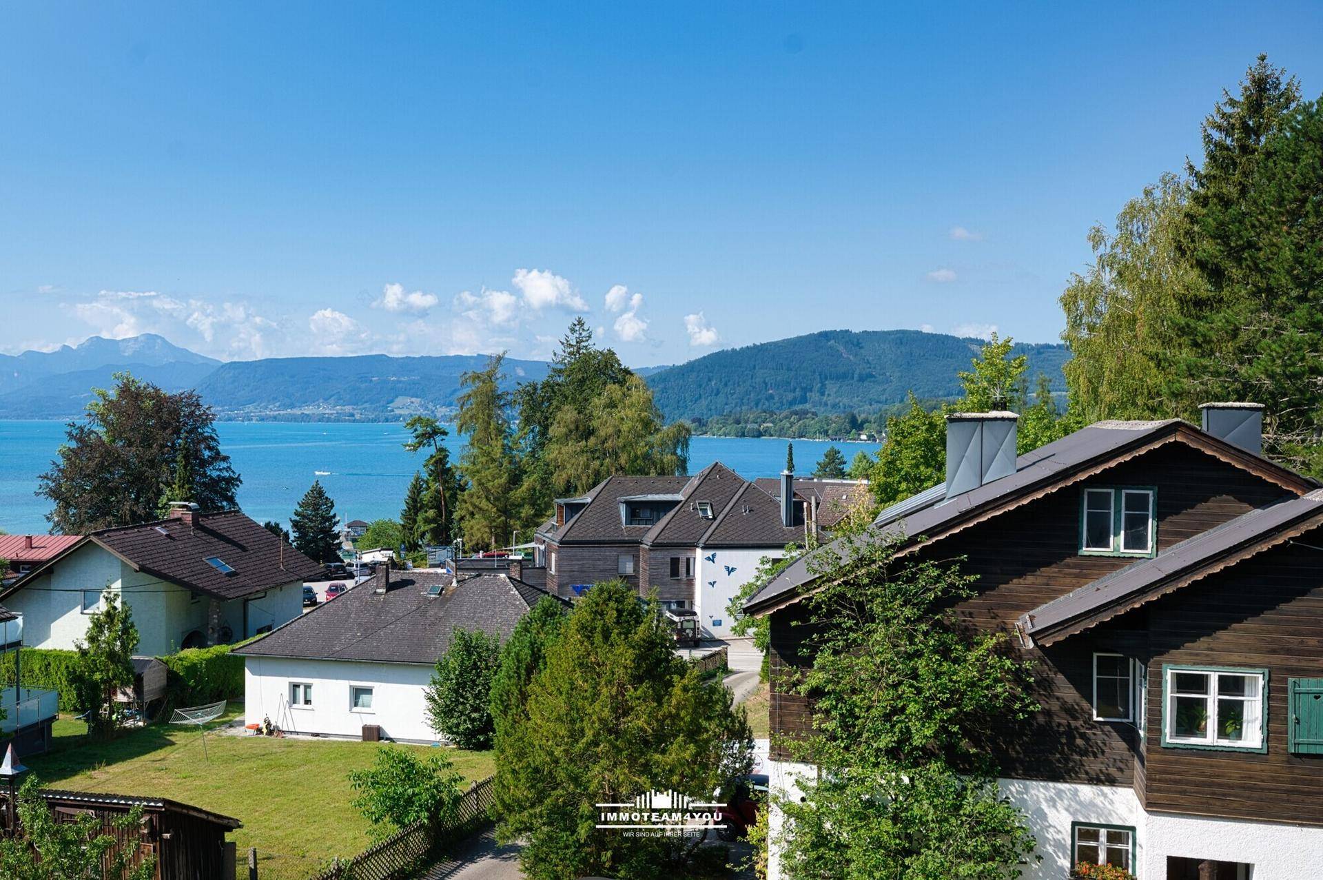 Weitläufiger Blick über die Dächer der Häuser auf den tiefblauen See und die bewaldeten Hügel.