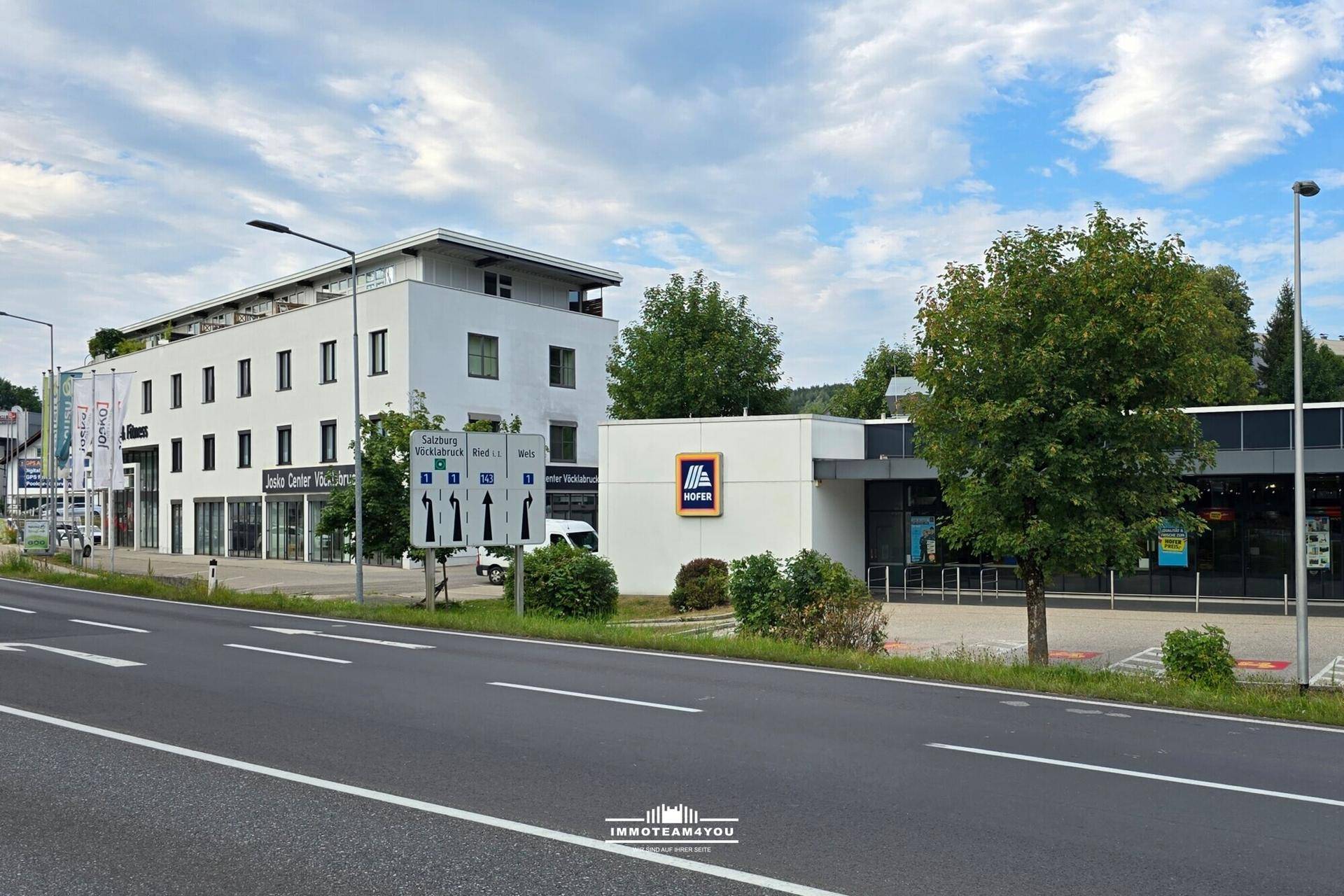 Modernes Geschäftsgebäude neben einem Supermarkt, umgeben von Bäumen unter einem bewölkten Sommerhimmel.