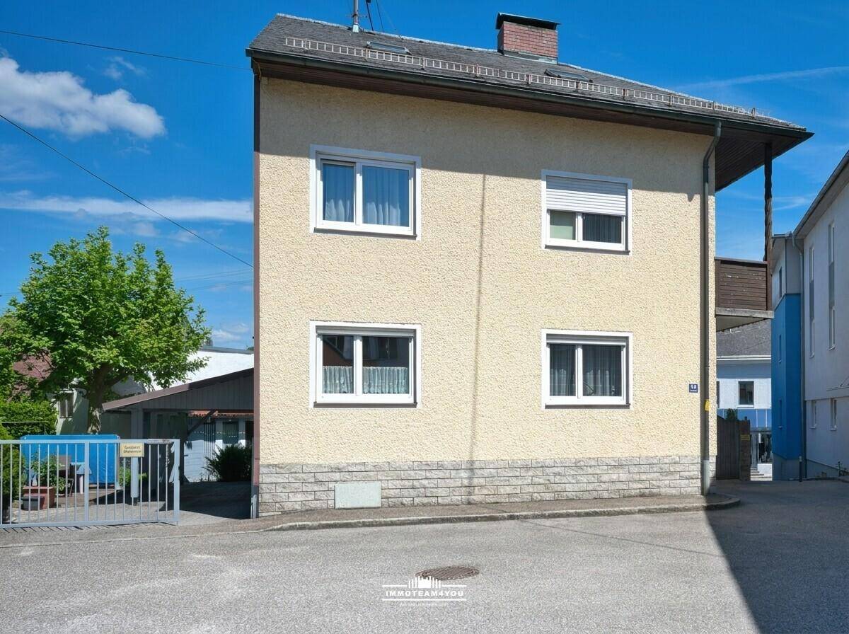 Vorderansicht des Hauses mit Putzfassade, Fenstern und Rollläden unter blauem Himmel.