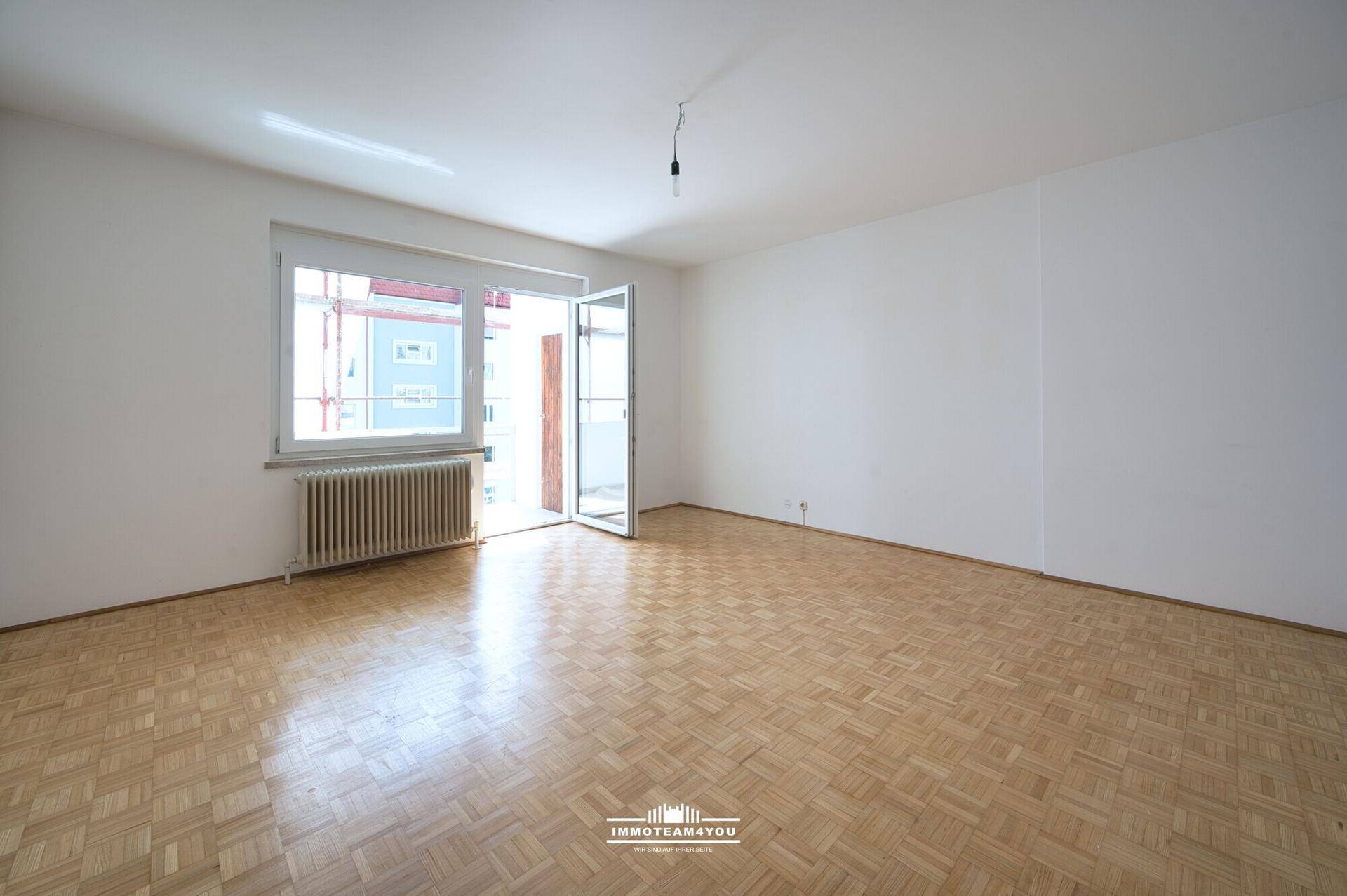 Helles, leeres Zimmer mit Fischgrätparkett, großem Fenster und Zugang zu einem Balkon.