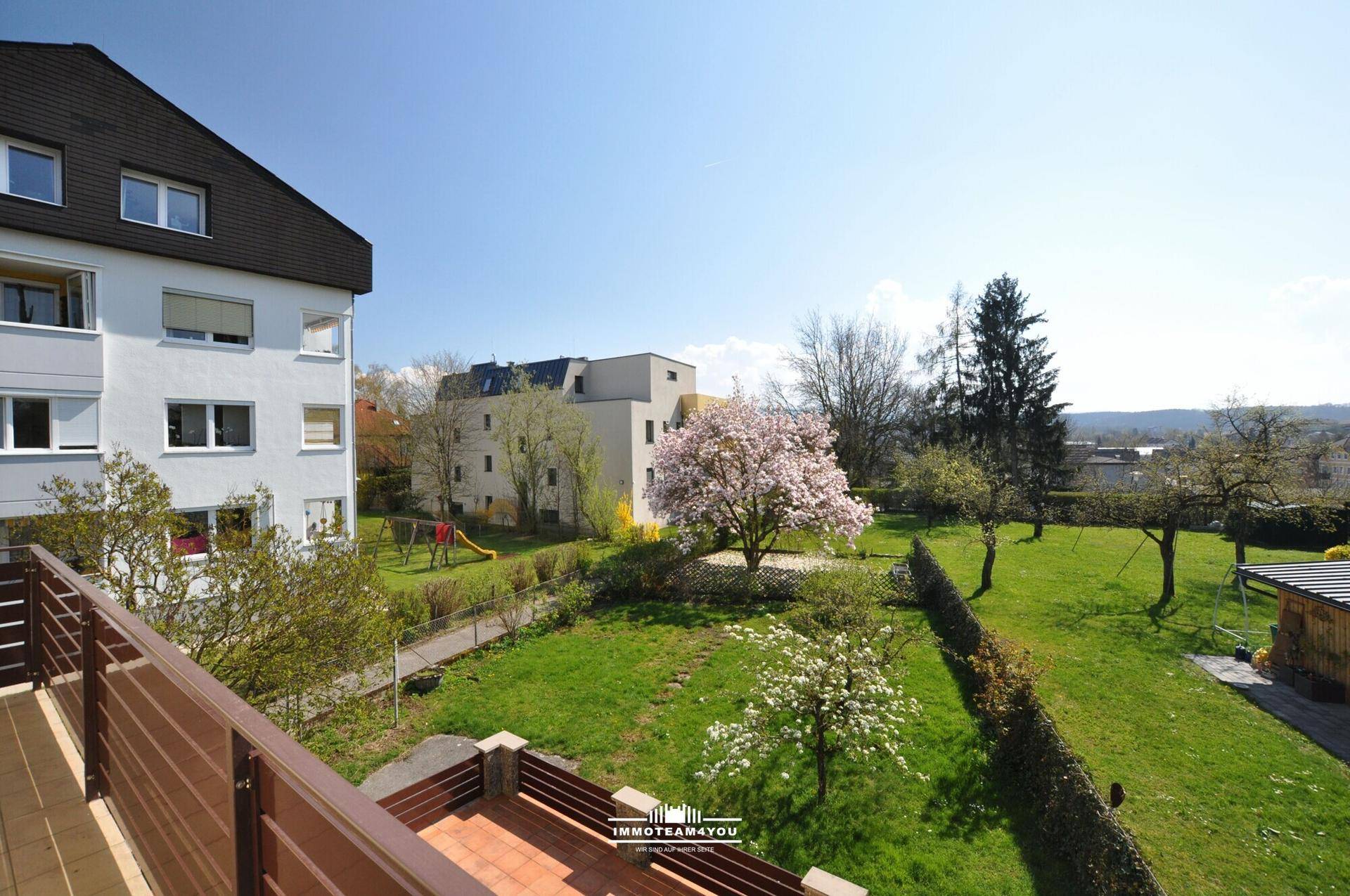 Weitläufiger Gartenbereich mit blühenden Bäumen und angrenzenden Gebäuden, gesehen vom Balkon.