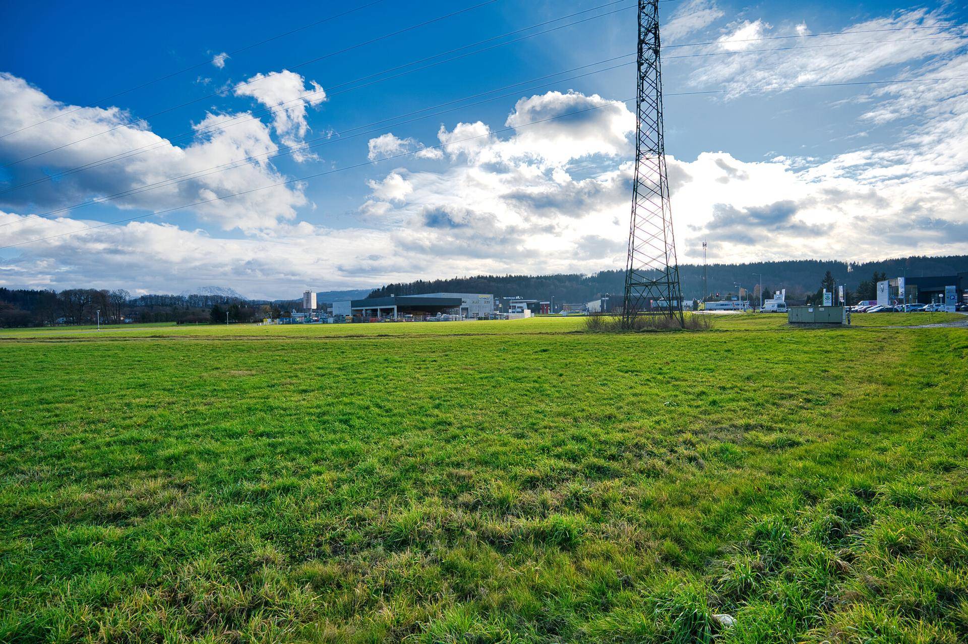 Weitläufige grüne Wiese mit einem hohen Strommast und Gebäuden in der Ferne unter blauem Himmel.