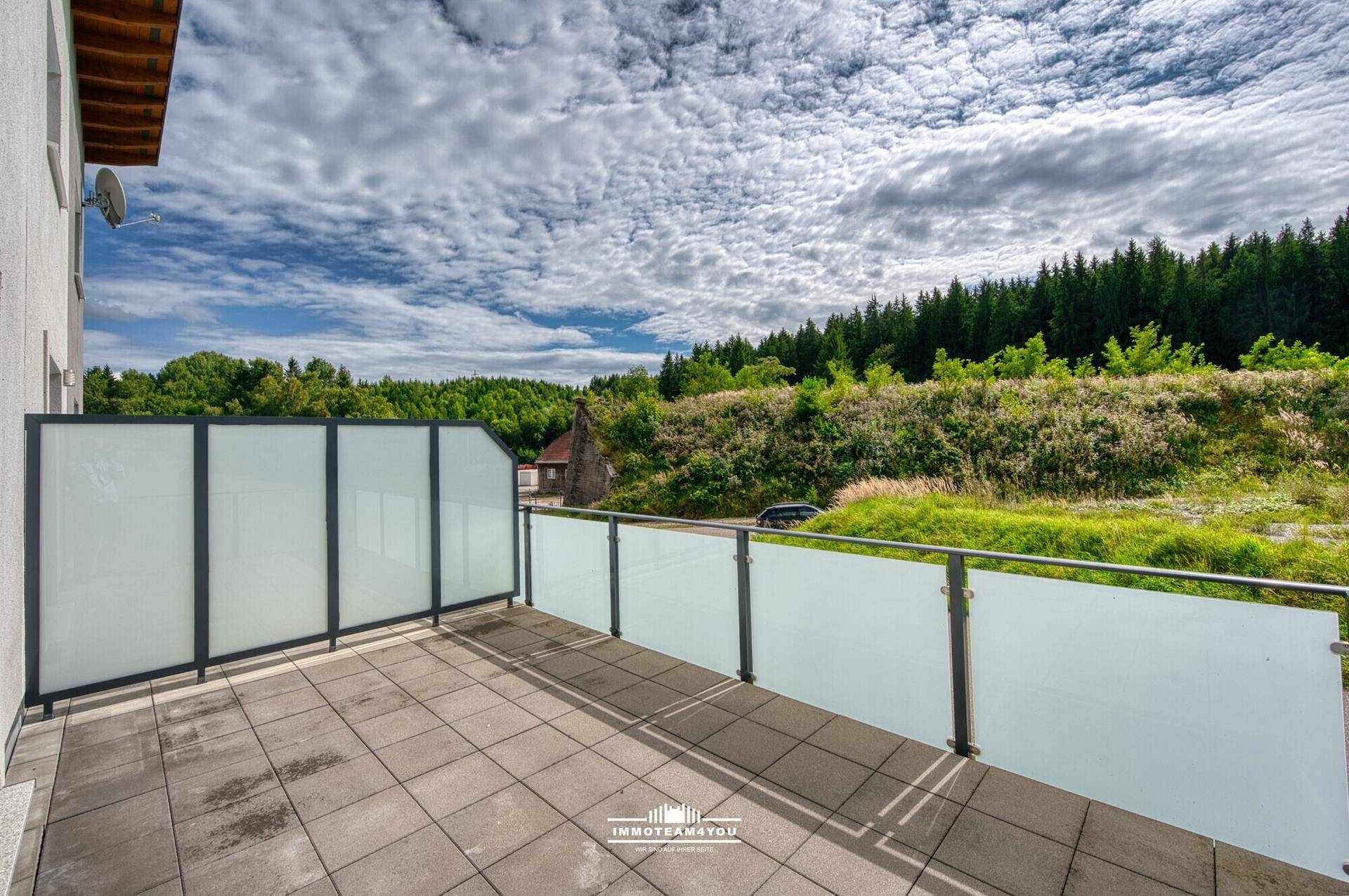 Geräumige Terrasse mit Glasgeländer und weitem Blick auf die grüne Landschaft und Wälder.
