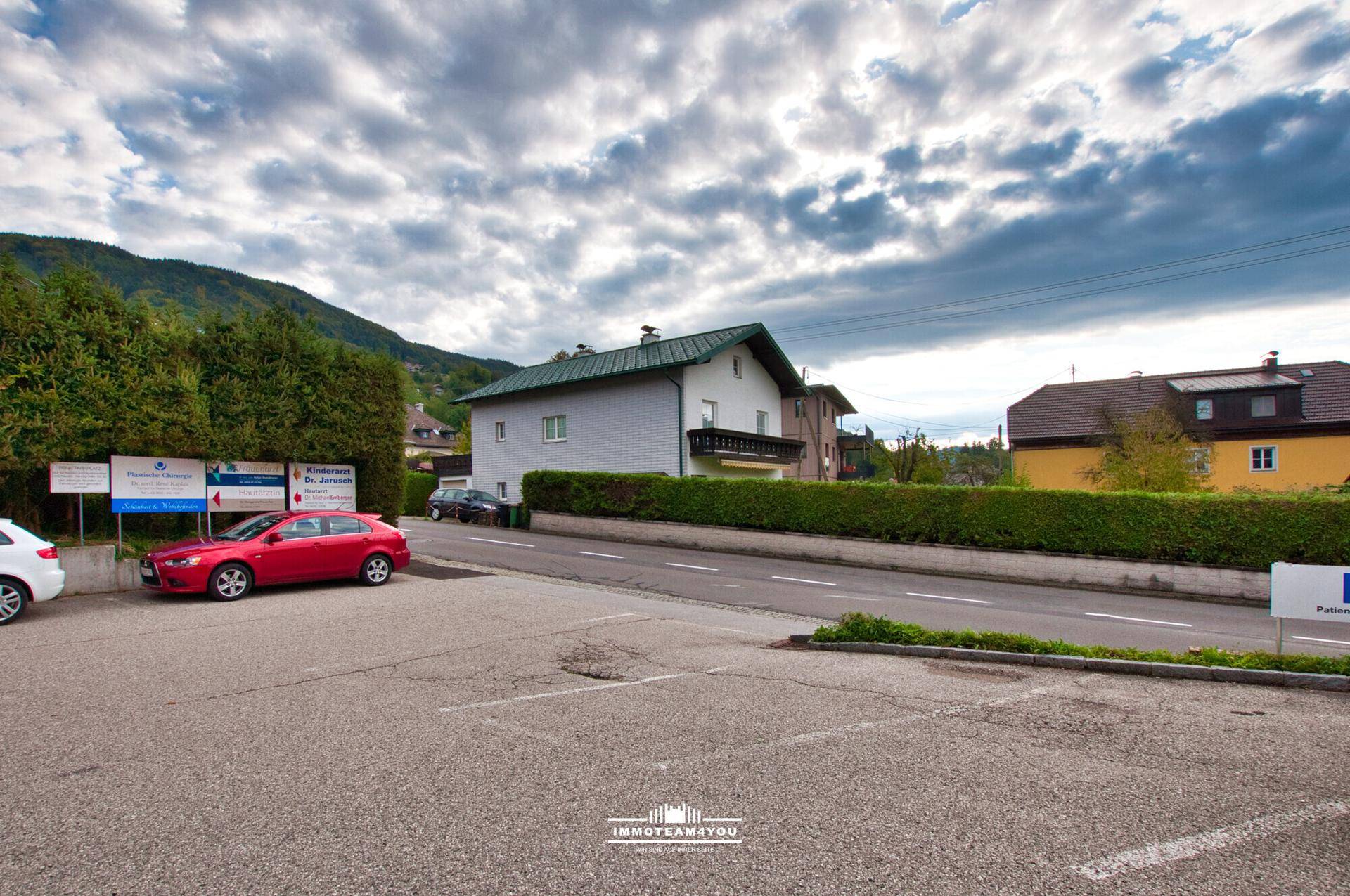 Außenansicht eines Gebäudes mit Parkplatz, umgeben von Bäumen und Bergen im Hintergrund.