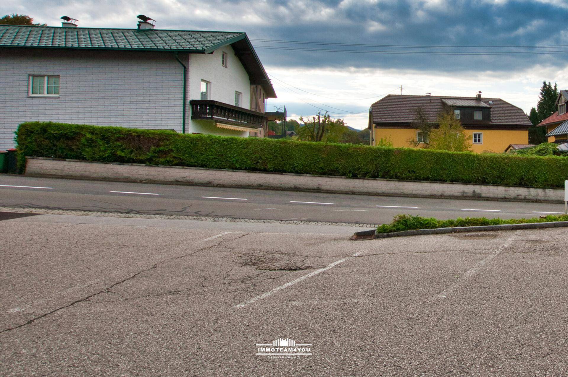 Parkplatz mit Blick auf eine Straße, Hecke und Wohnhäuser in ländlicher Umgebung unter bewölktem Himmel.