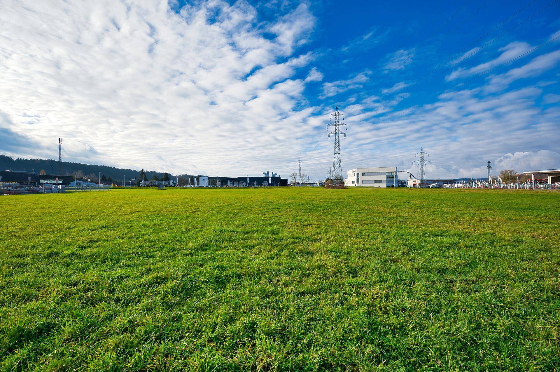 Grüne Wiese mit einem Strommast und Gebäuden in der Ferne unter einem bewölkten Himmel.