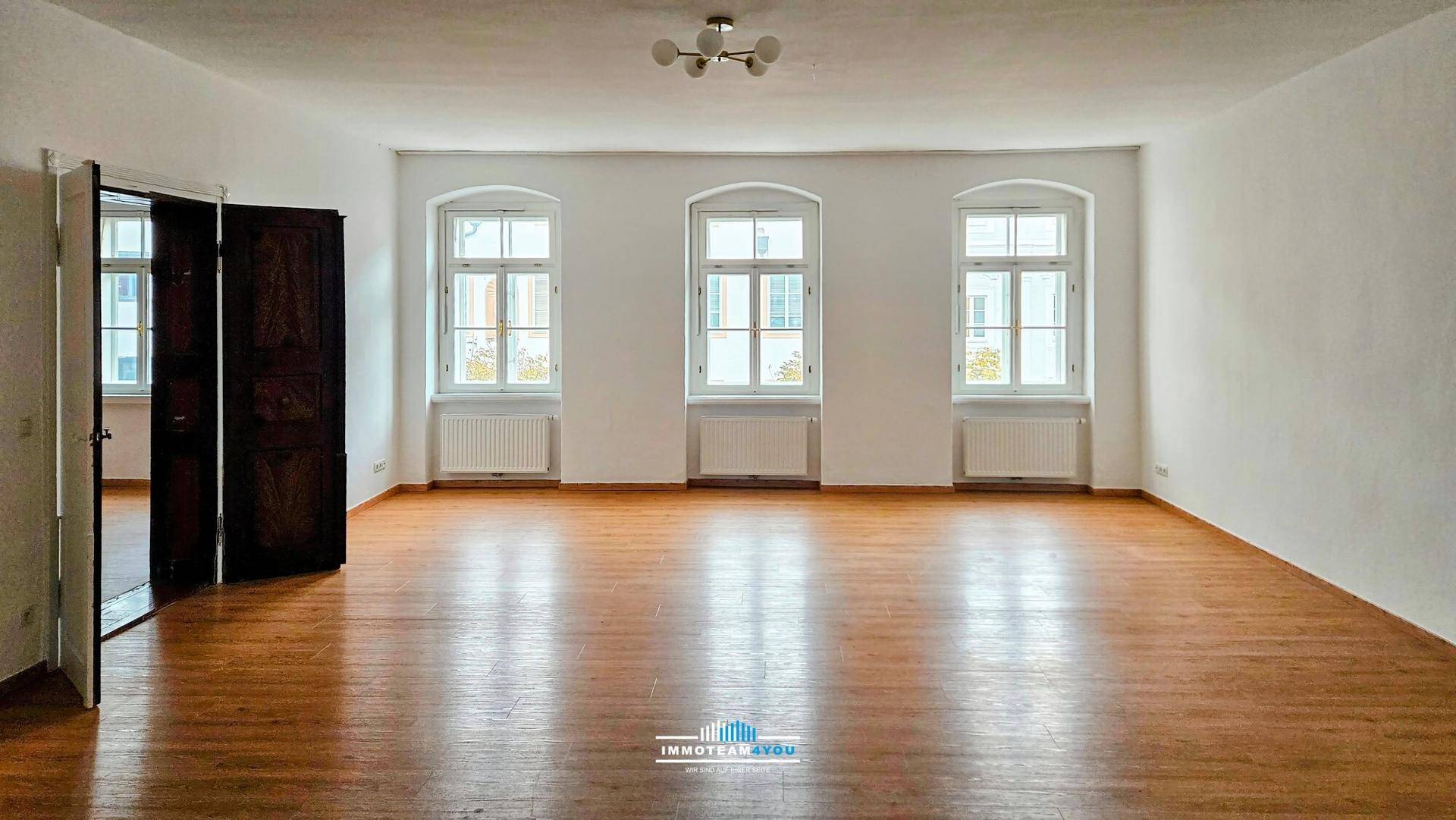 Helles, leeres Zimmer mit Holzboden, hohen Decken und drei großen Fenstern.