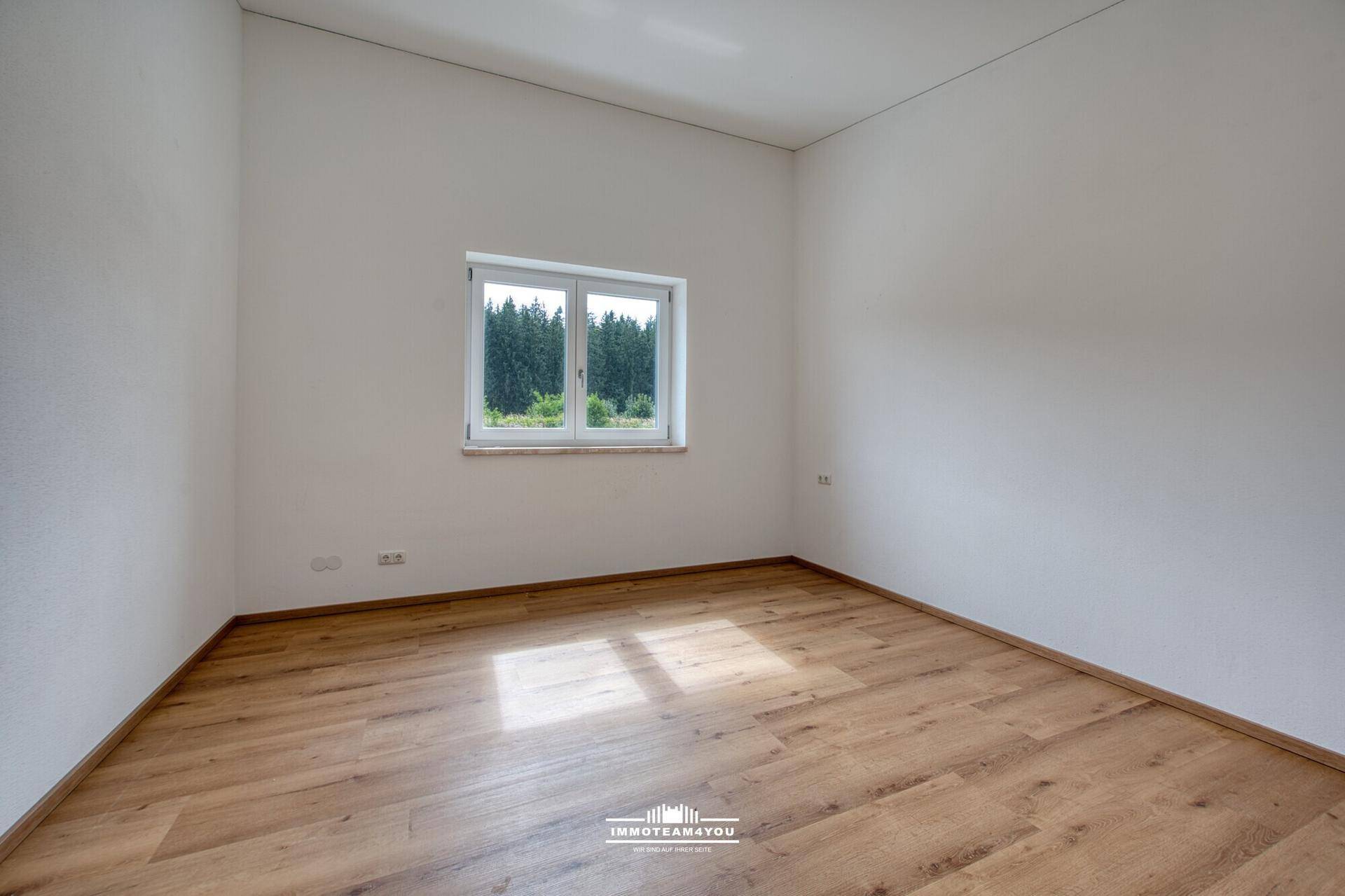 Leeres Schlafzimmer mit Holzboden, weißen Wänden und Fenster mit Blick ins Grüne.