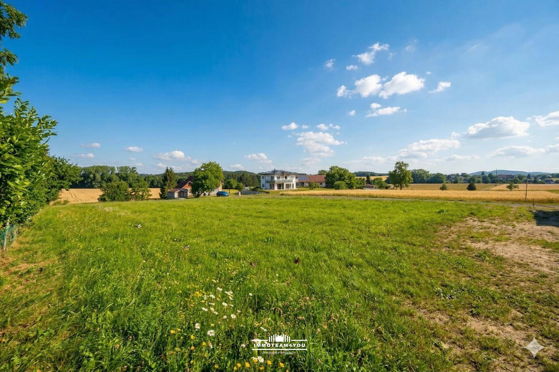 Weite grüne Wiesen mit vereinzelten Häusern und Feldern in der Ferne unter blauem Himmel.