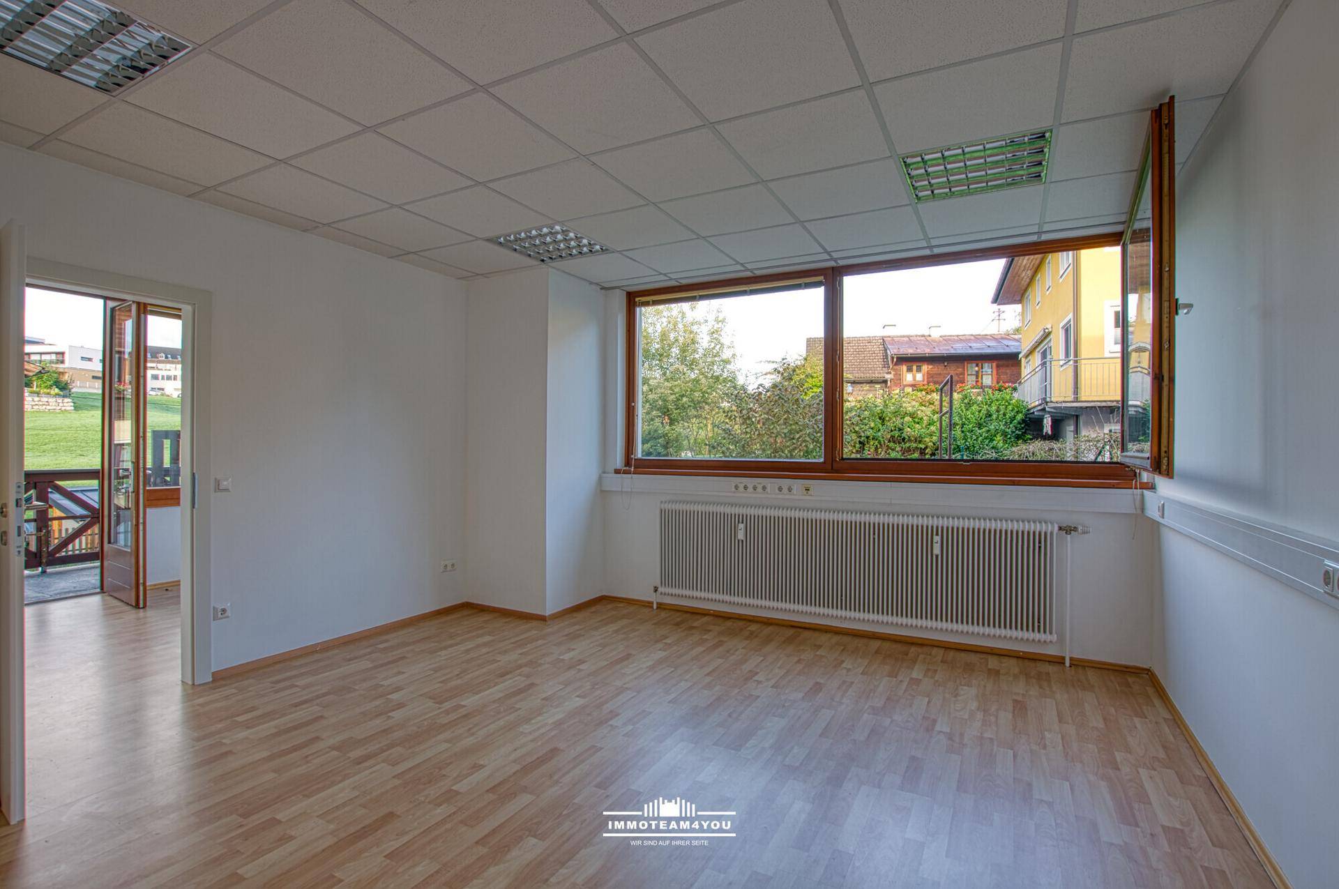 Heller Büroraum mit Laminatboden, großen Fenstern und Zugang zu einem Außenbereich.