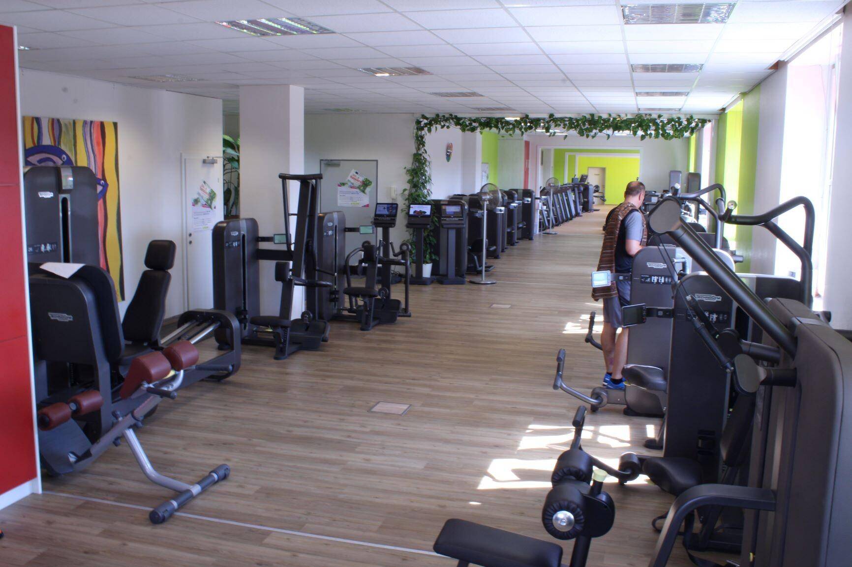 Langer Fitnessraum mit einer Vielzahl von Krafttrainingsgeräten und hellem Holzboden.