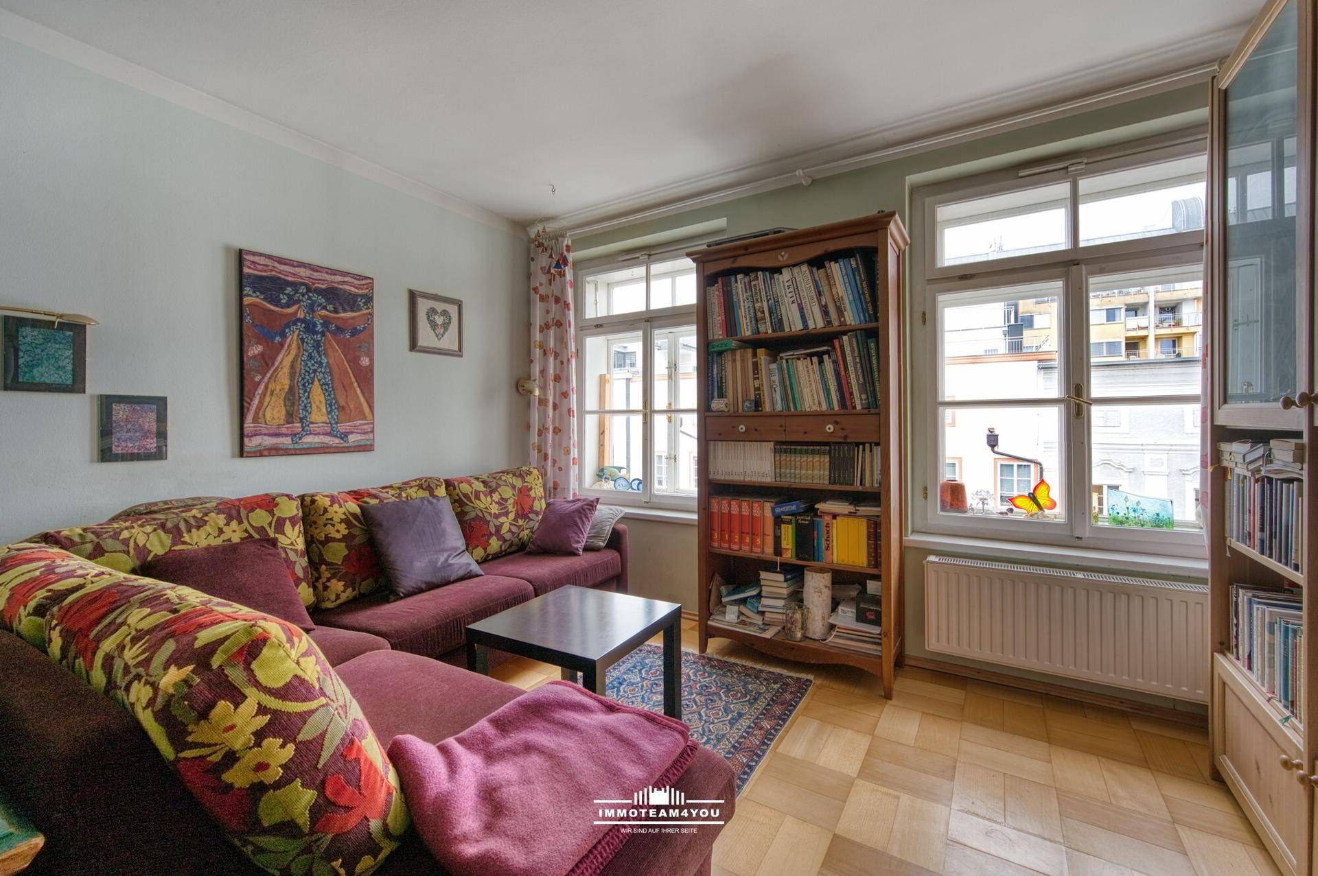 Gemütliches Wohnzimmer mit farbenfrohem Sofa, Bücherregal und Blick auf die Stadt durch die Fenster.