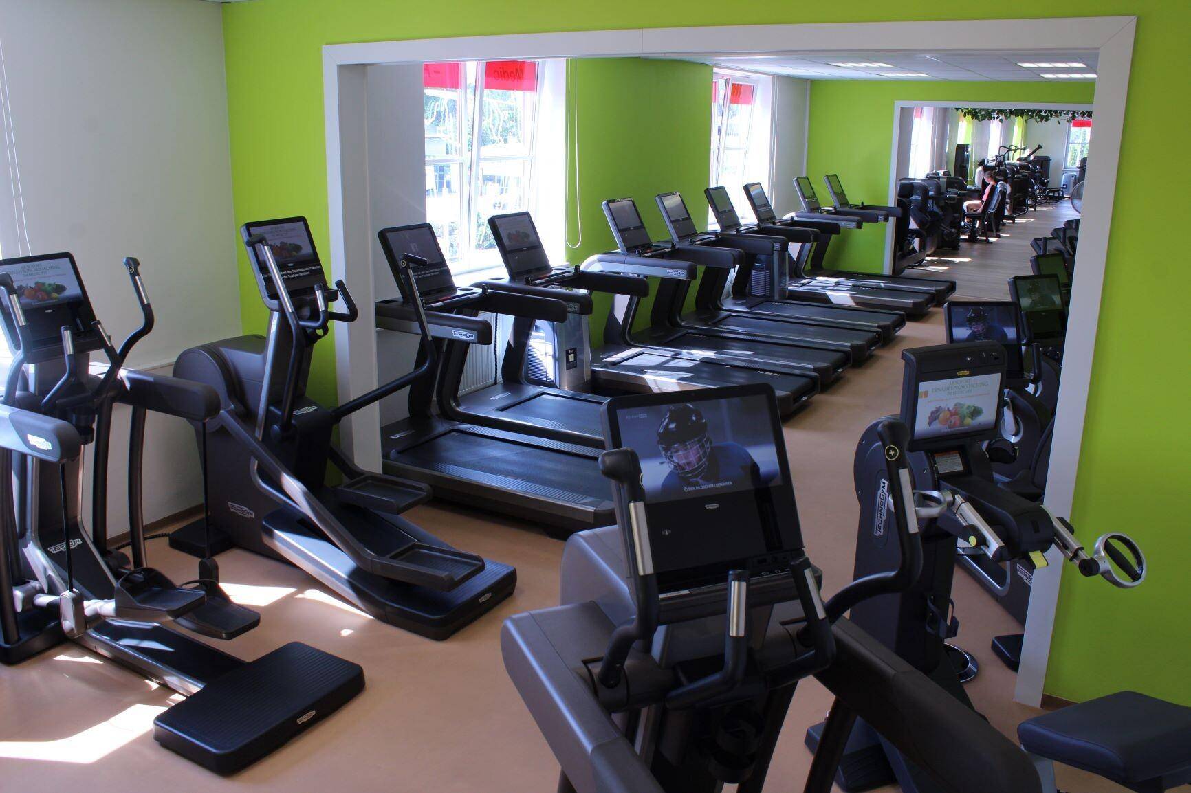 Heller Fitnessraum mit vielen Cardiogeräten und einem großen Wandspiegel, ideal für Ausdauertraining.