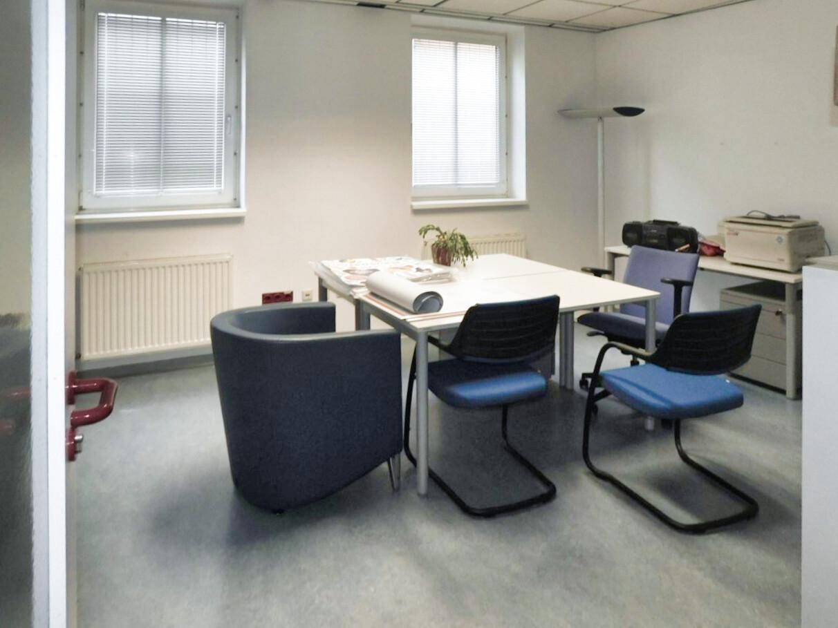 Besprechungsraum mit zwei Fenstern, einem Tisch und verschiedenen Stühlen, ausgestattet für Teamarbeit.