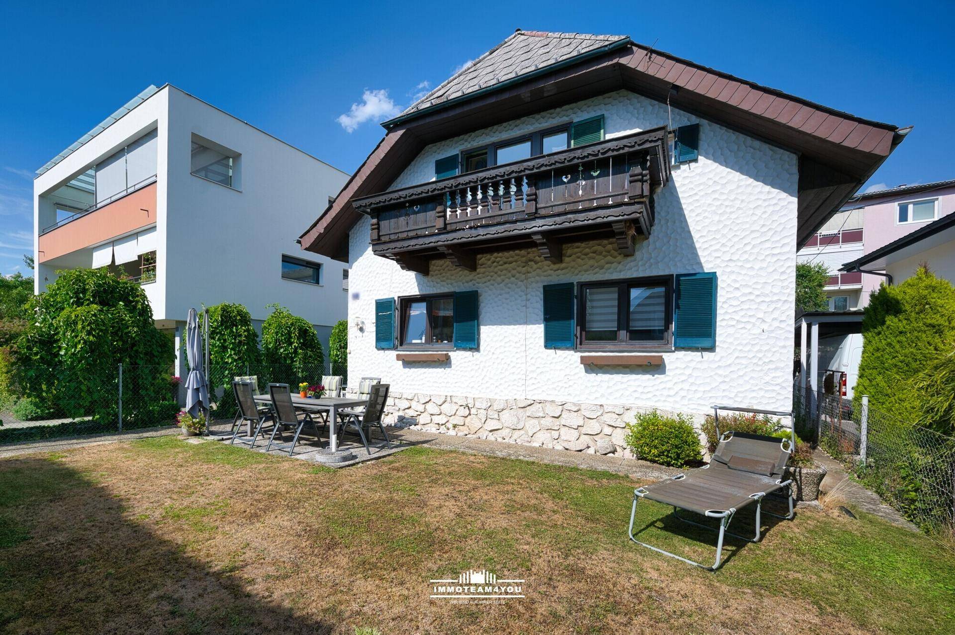 Sonniger Garten mit Terrasse und Liegestuhl, umgeben von grünen Büschen und Nachbargebäuden.