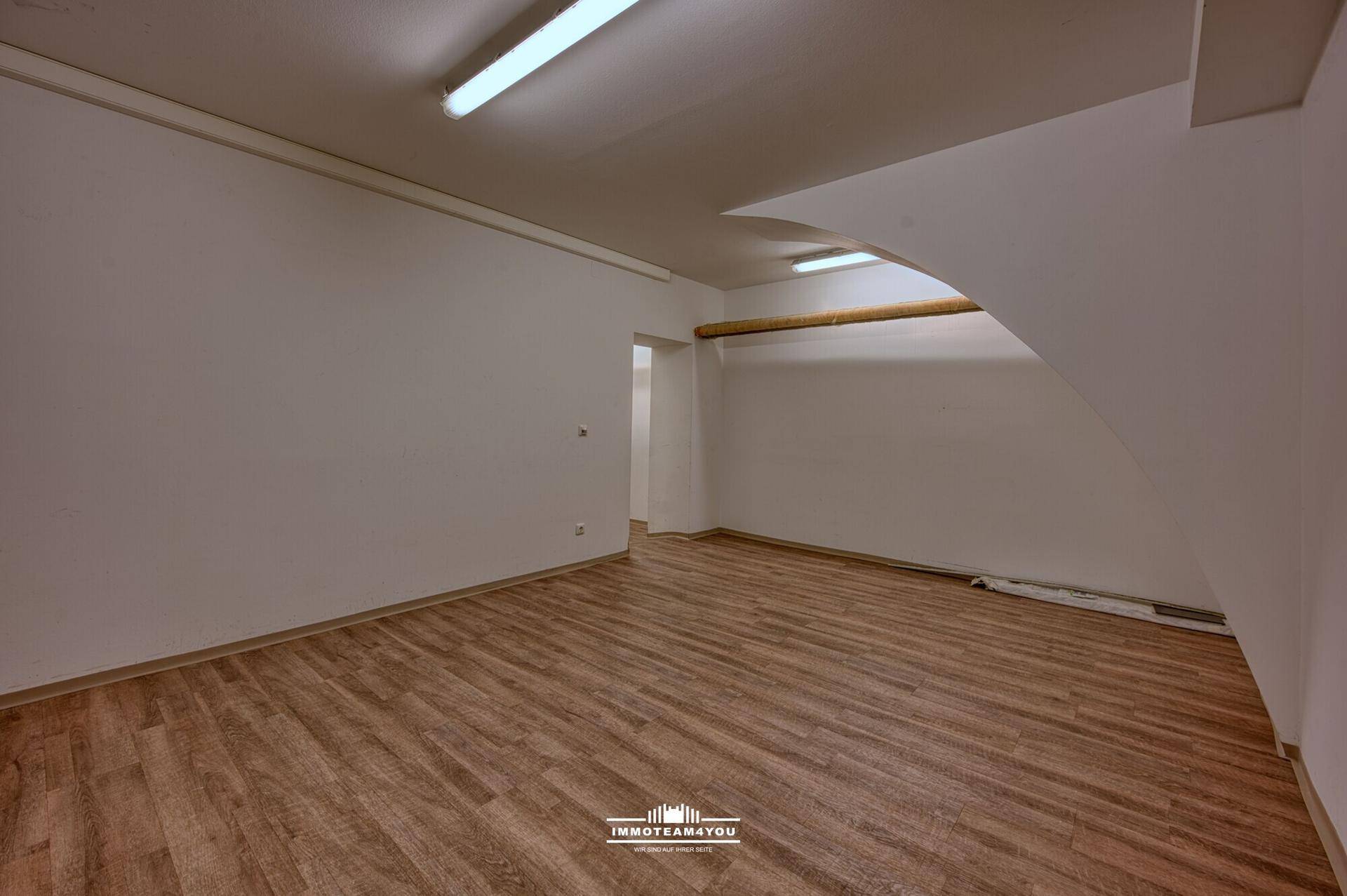 Leerer Lagerraum mit Laminatboden, weißen Wänden und einer leicht schrägen Decke im hinteren Bereich.