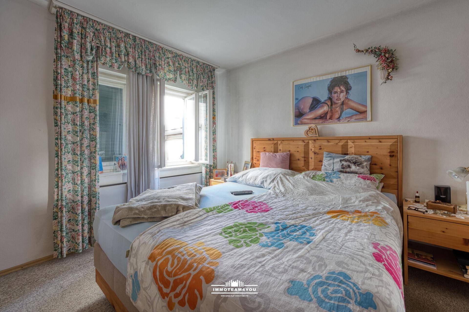 Schlafzimmer mit Doppelbett, Holzmöbeln und floralen Vorhängen, hell durch große Fenster.