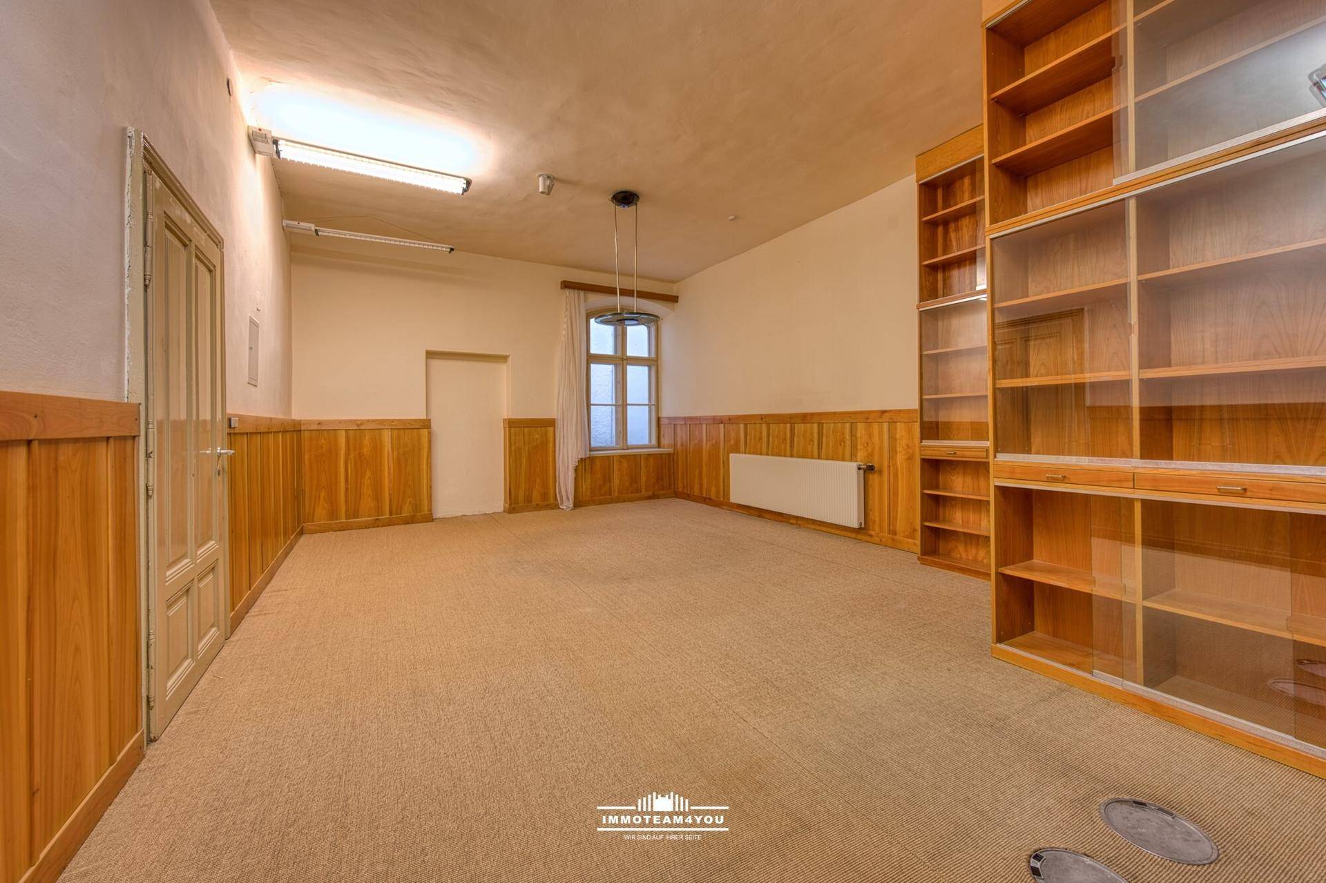 Geräumiger Büroraum mit beigem Teppichboden, Holzvertäfelung und großen, eingebauten Bücherregalen, ideal für Archivierung.