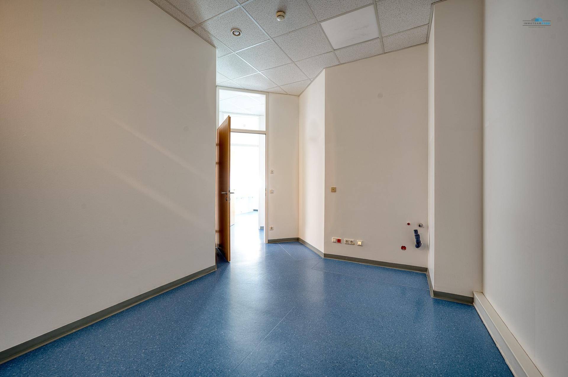 Leerer Raum mit blauer Bodenfläche, weißen Wänden und sichtbaren Wasseranschlüssen für eine mögliche Nutzung.