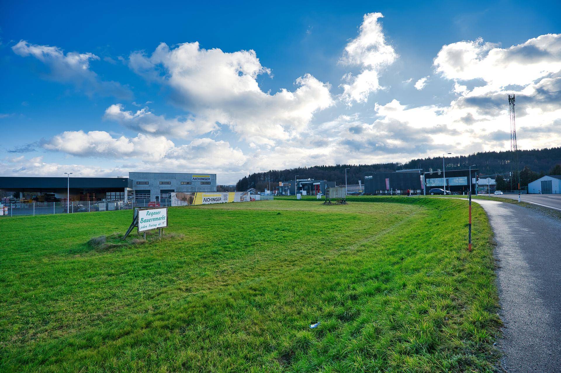 Grüne Wiese mit einem Hinweisschild und Gebäuden in der Ferne unter einem bewölkten Himmel, der die Landschaft beleuchtet.
