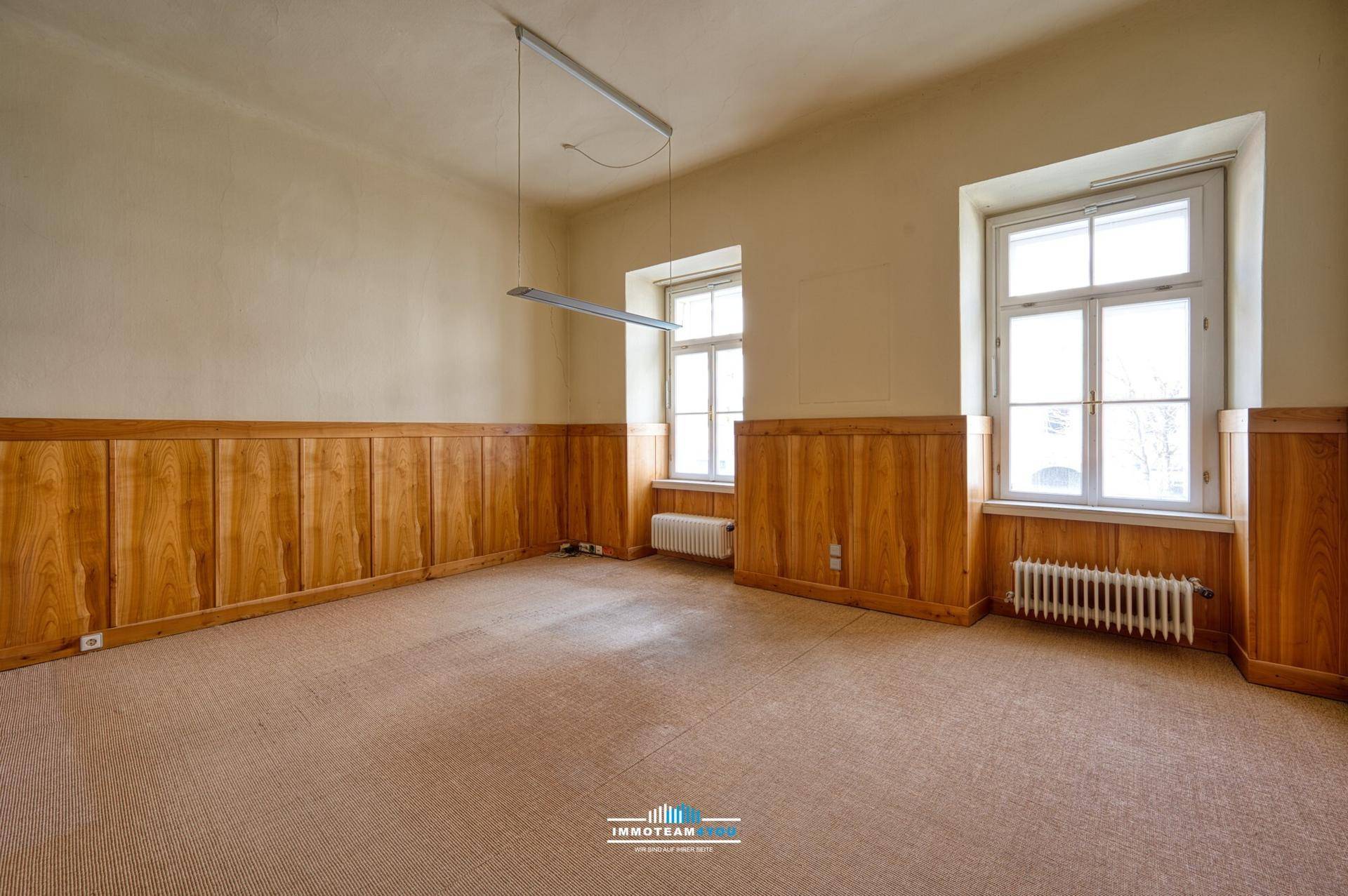 Geräumiges Zimmer mit Holzvertäfelung an den Wänden, Teppichboden und großen Fenstern.