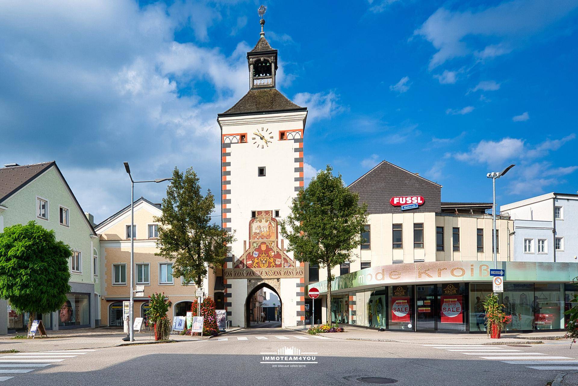 Historischer Stadtturm mit Uhr und Malereien, umgeben von Geschäften und Gebäuden an einem sonnigen Tag.