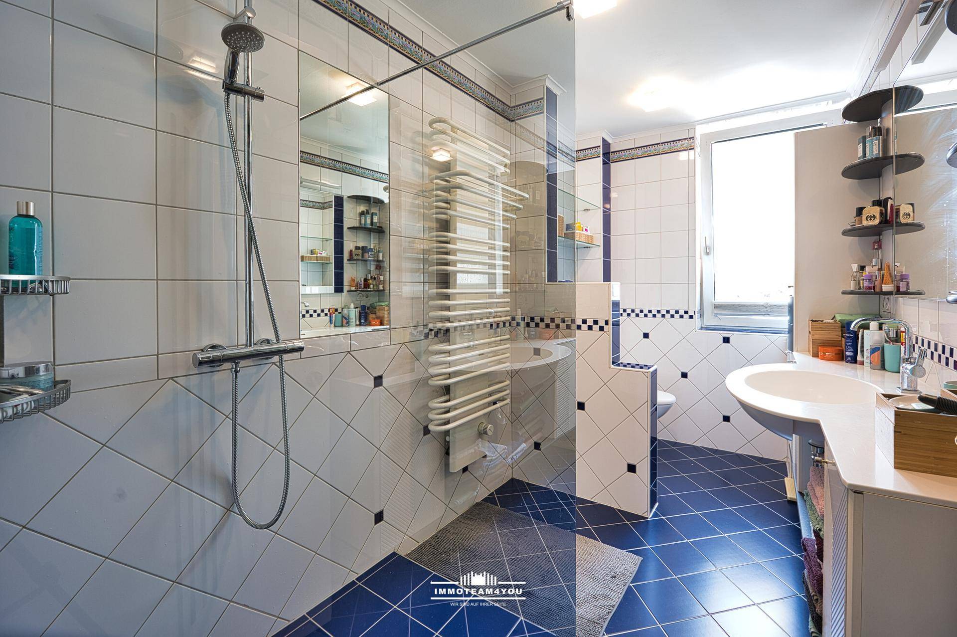 Helles Badezimmer mit blauen und weißen Fliesen, Dusche, Fenster und beheiztem Handtuchhalter.