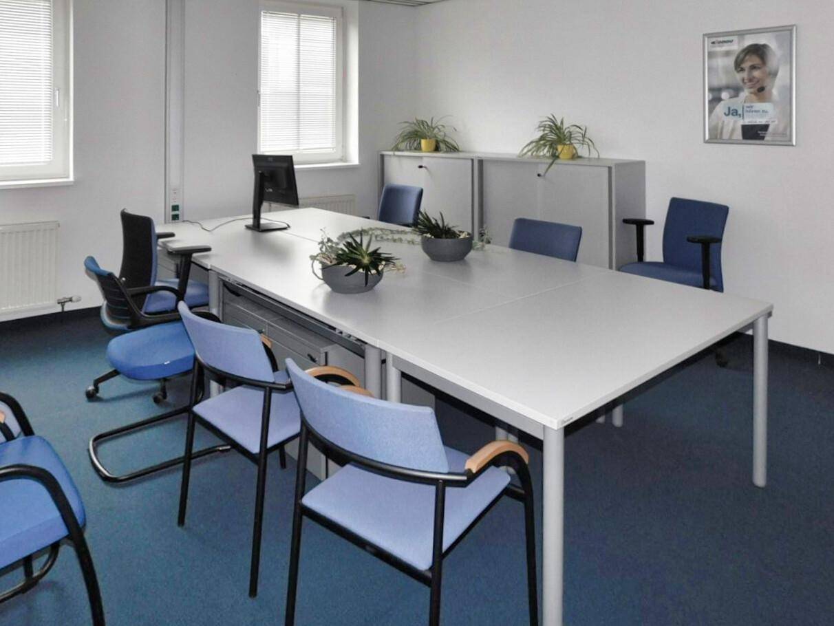 Großer Besprechungsraum mit langem Tisch, mehreren Stühlen und zwei Fenstern für natürliches Licht.