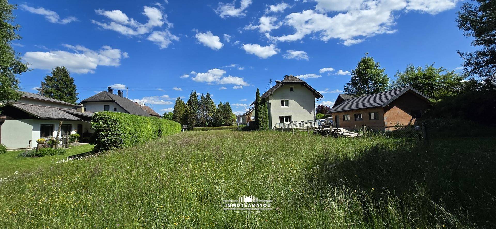 Große, überwachsene Rasenfläche mit mehreren Wohnhäusern und Bäumen unter blauem Himmel.