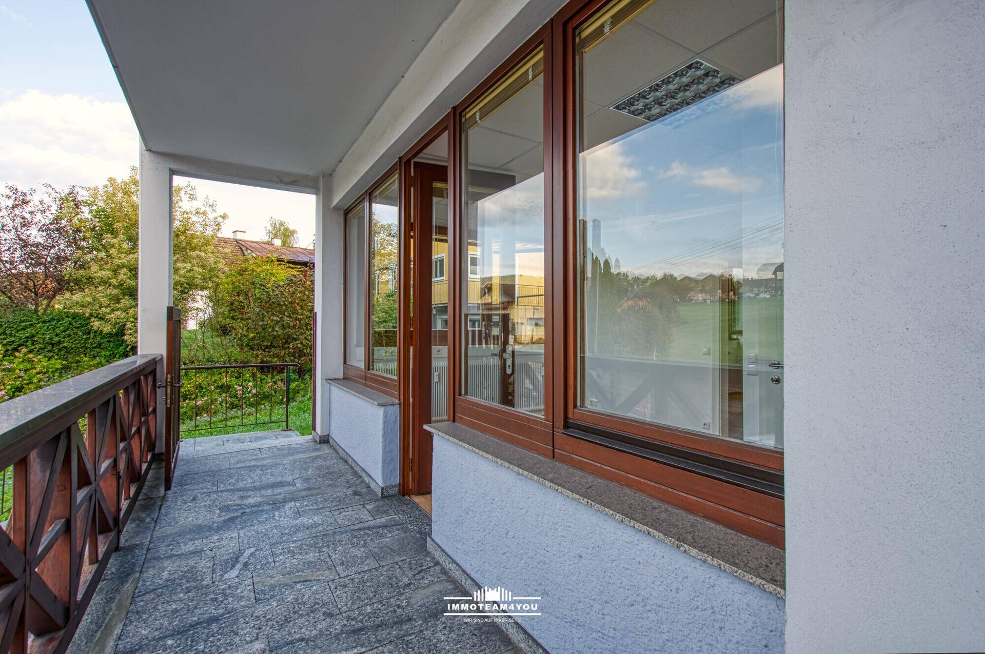 Geräumiger Balkon mit Steinboden und Holzgeländer, umgeben von grüner Landschaft und Bäumen.