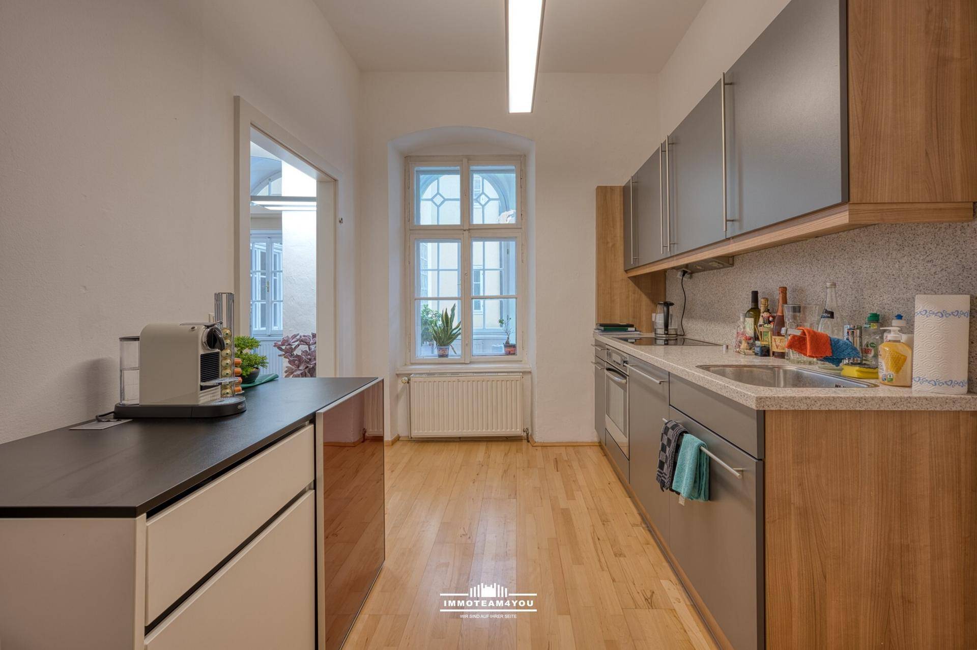 Helle Küche mit Holz- und grauen Schränken, Holzboden und einem Fenster zum Innenhof.