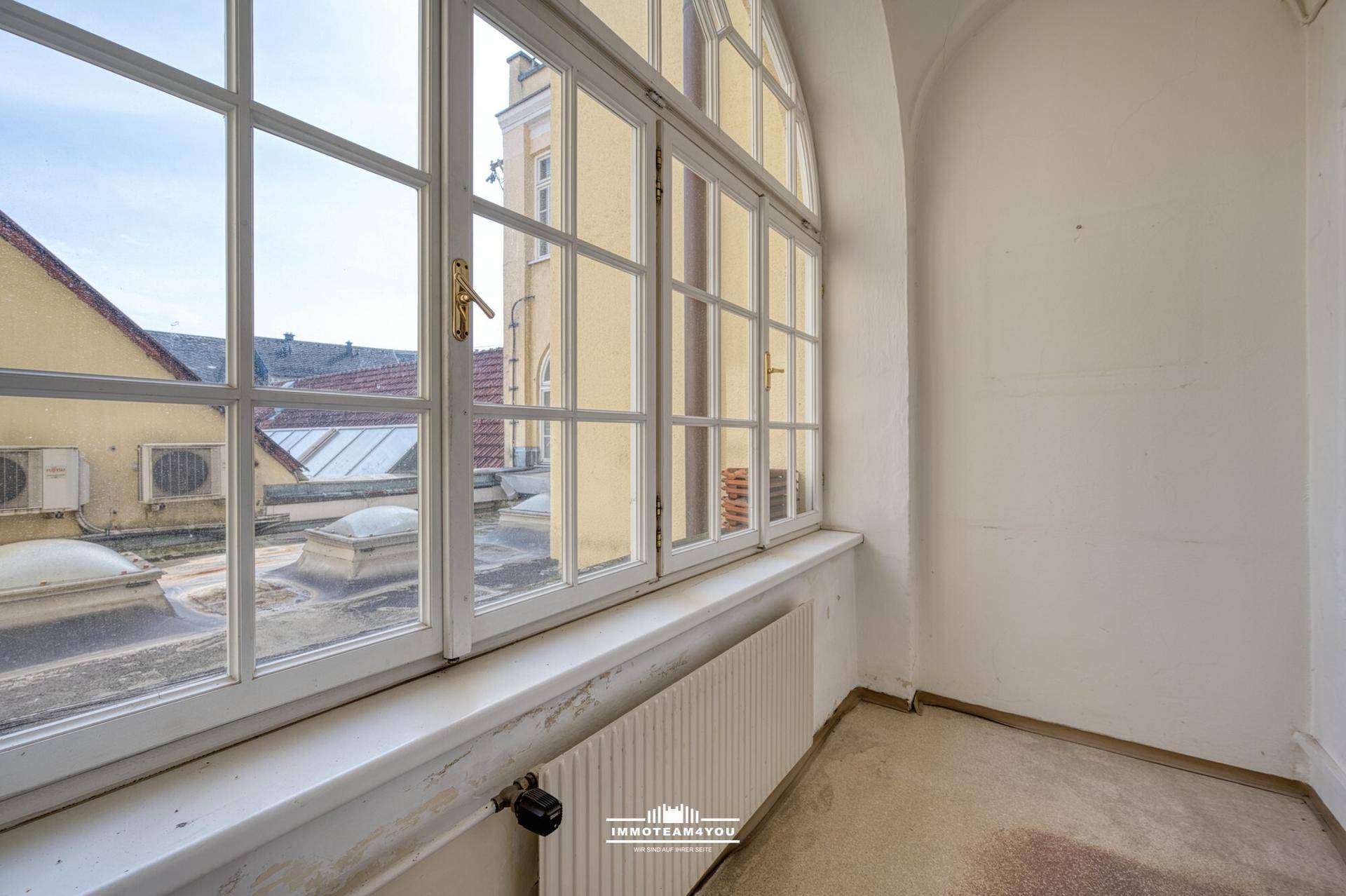 Kleiner Raum mit großem Bogenfenster und Blick auf Dächer, mit Heizkörper und einfacher Ausstattung.