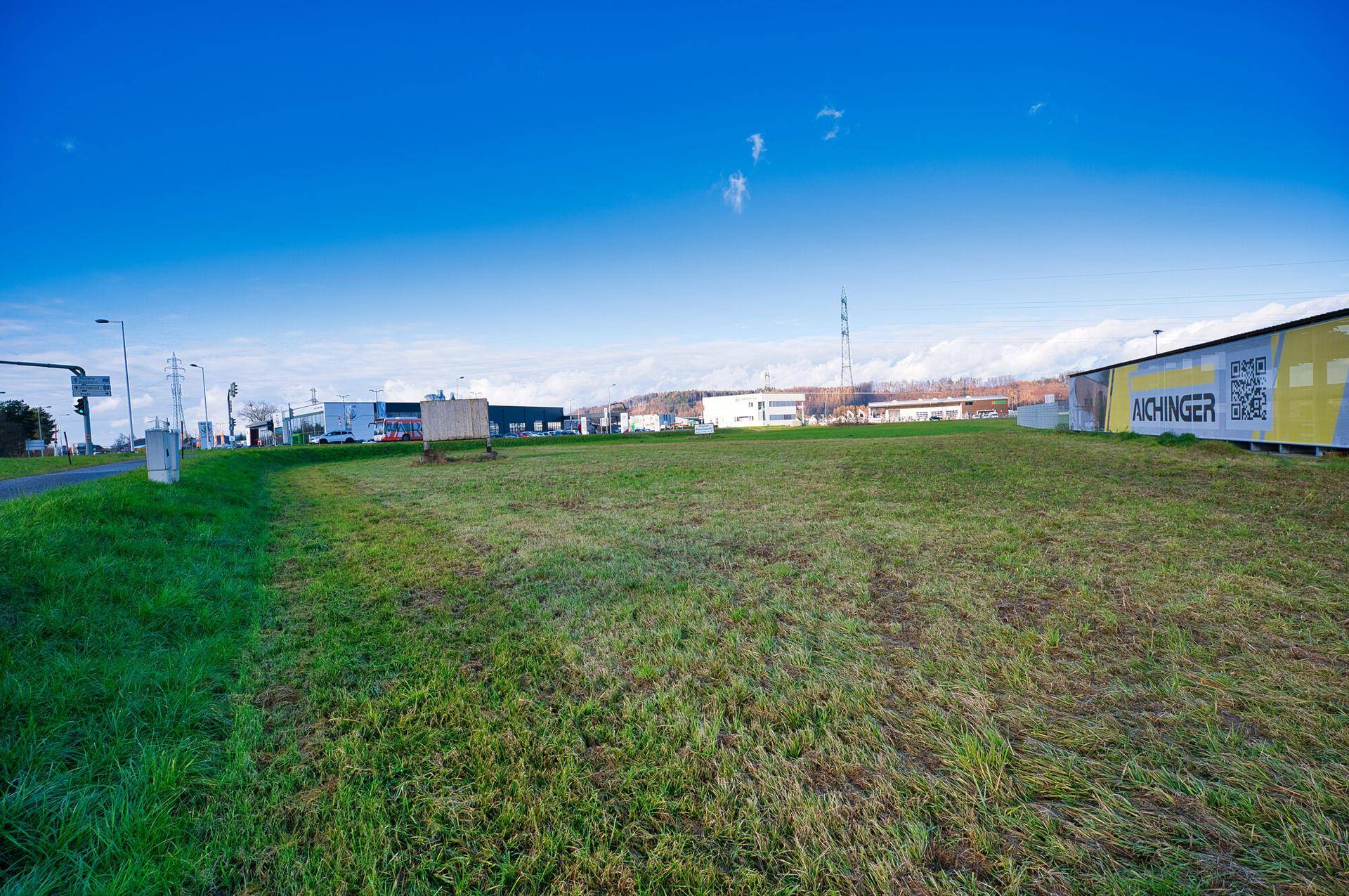 Grüne Grasfläche neben einer Straße mit Werbeschildern und Gebäuden im Hintergrund unter blauem Himmel.