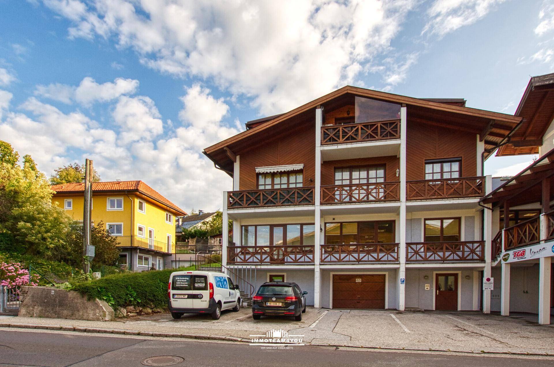 Mehrstöckiges Gebäude mit Holzfassade, Balkonen und Parkplätzen vor dem Eingangsbereich.