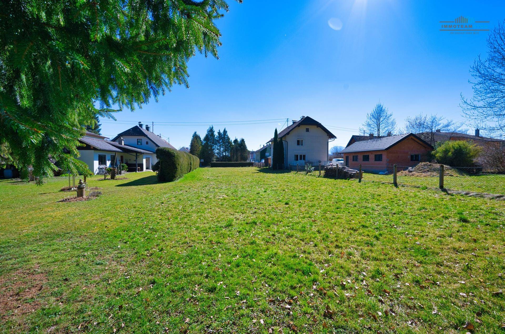 Sonnige Rasenfläche mit mehreren Wohnhäusern und Bäumen in der Ferne unter klarem Himmel.