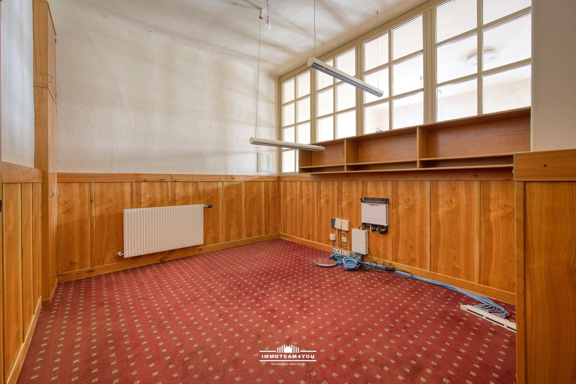 Heller Büroraum mit rotem Teppichboden, Holzvertäfelung und großen Fenstern, ausgestattet mit eingebauten Regalen.