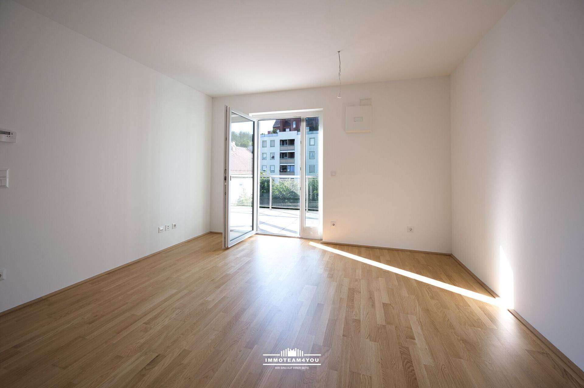 Heller, leerer Raum mit Holzboden und Zugang zu einem Balkon, der einen Blick auf Gebäude und Bäume bietet.