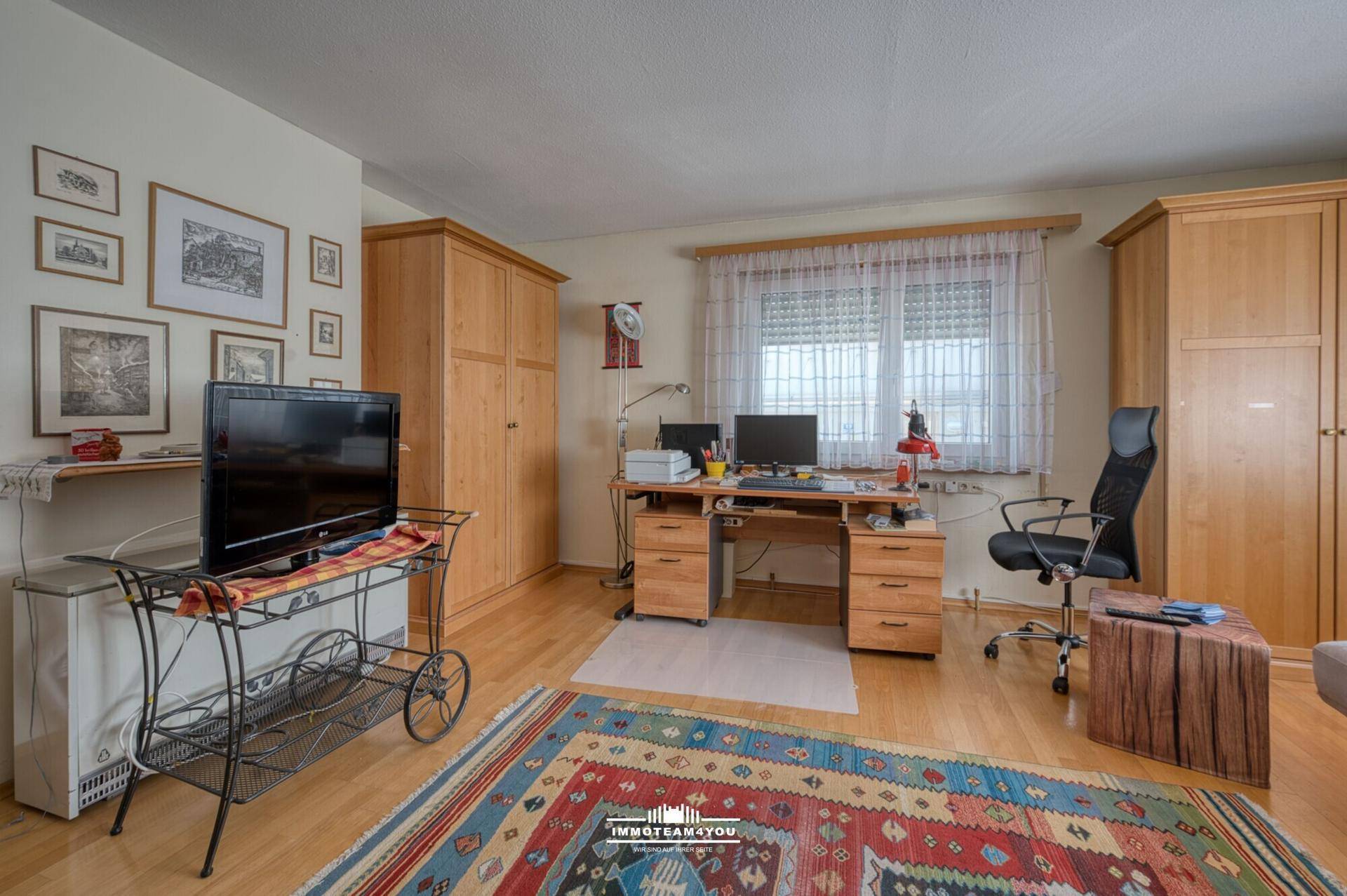 Helles Arbeitszimmer mit Holzmöbeln, Schreibtisch und einem farbenfrohen Teppich.