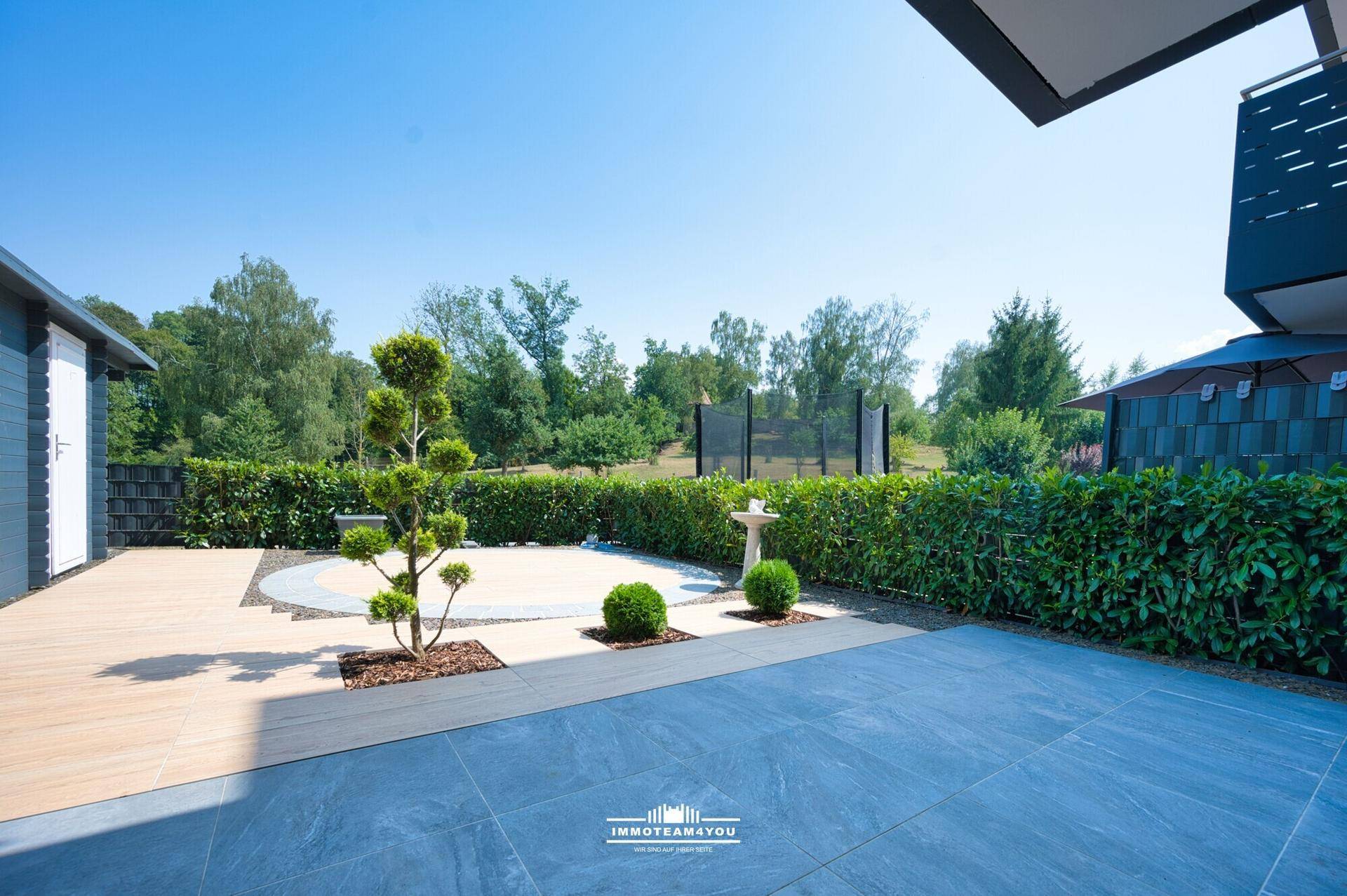 Moderne Terrasse mit verschiedenen Bodenbelägen, umgeben von Hecken und Bäumen.