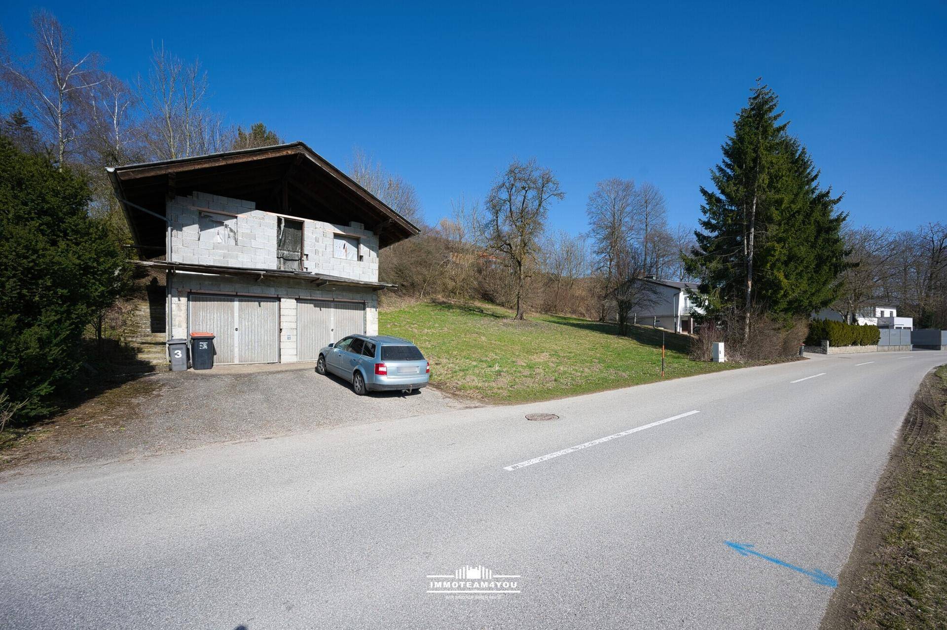 Haus im Bauzustand mit Garagen und einem Auto an einer Straße, umgeben von Grünflächen.