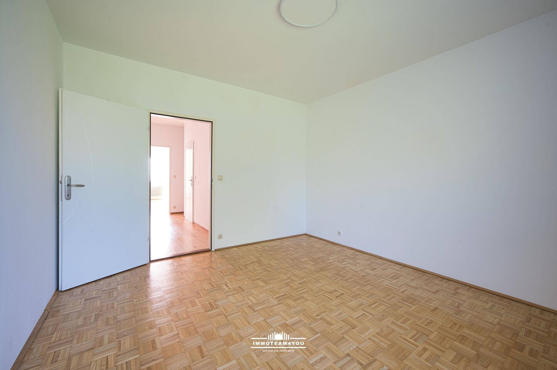 Geräumiges Zimmer mit Parkettboden und offener Tür, die den Blick in den Flur freigibt.